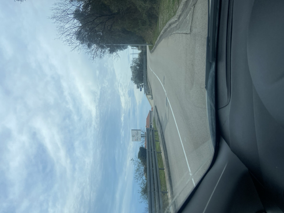 A road ascends slightly, marked with a central white line. Guardrails flank the roadside, and a sign indicating "Oliva Villages" is visible. The sky is blue, adorned with streaky clouds. Trees border the opposite side of the road. The photograph appears to be taken from within a car. The vehicle's dashboard and interior are partially visible in the foreground.