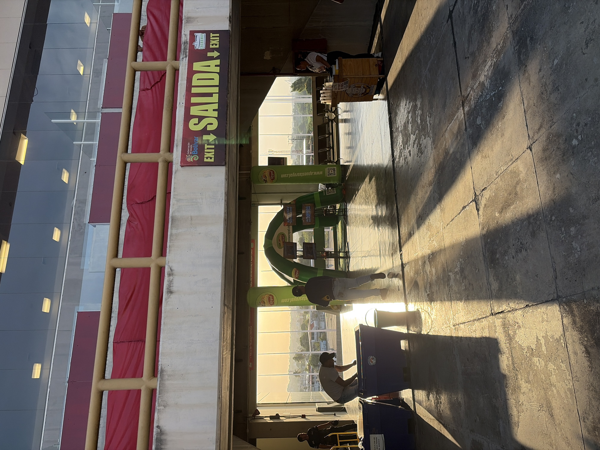 The image shows an exterior view of an exit point at a location in Ciudad Guayana, Venezuela. The scene is characterized by strong sunlight casting shadows on the paved ground. A red and gold-painted structure with glass panels is visible on the left, adjacent to an "EXIT SALIDA" sign. In the center, there is a green archway display and a table with cups, possibly for beverage sales. People are scattered throughout the area, including workers or event staff who are setting up or cleaning. The background features a landscape with trees and structures under a bright sky. The image appears to be taken in the late afternoon.