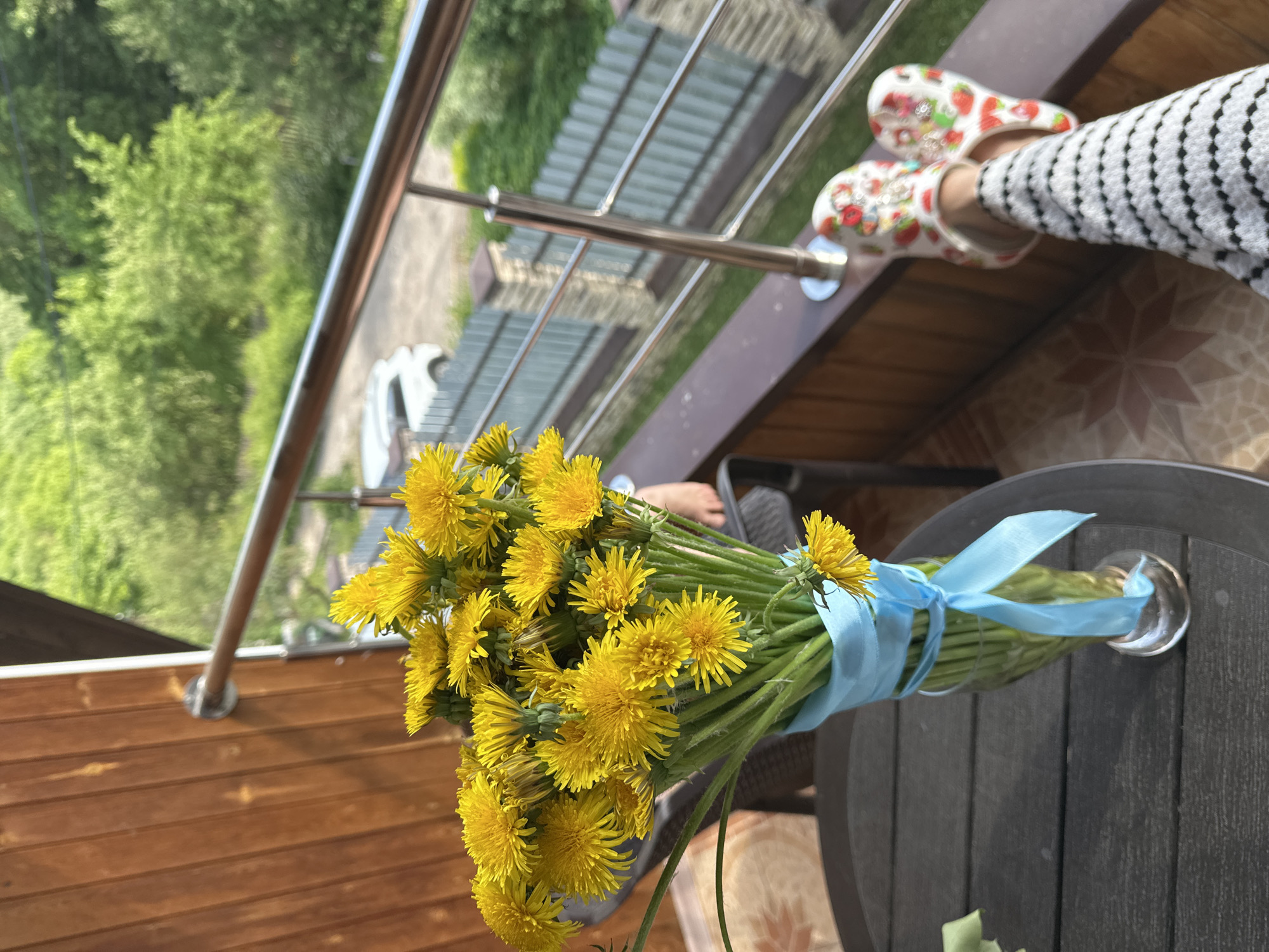 A close-up, high-angle shot captures a bouquet of dandelions tied with a blue ribbon, placed in a glass vase on a dark brown table, likely on a balcony. A person is present with feet visible wearing strawberry-patterned clogs and striped pants. The balcony features wooden paneling, a tiled floor, and a metal railing that allows a view of trees and a building with a gray roof. The perspective suggests the image was taken in Nizhny Novgorod, Russia.