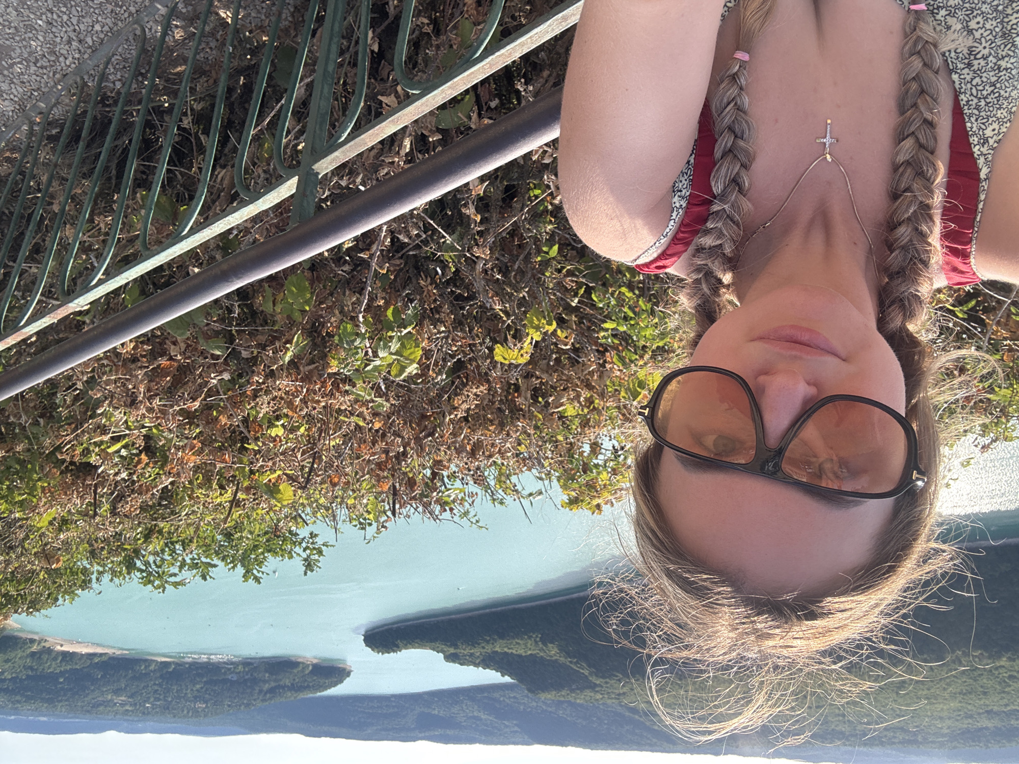 A young woman with blonde hair styled in two braids is taking a selfie outdoors. She is wearing large, dark-rimmed sunglasses with orange lenses, a light-colored dress with a floral pattern, and a red top with thin straps. A silver cross necklace hangs around her neck. The background features a body of water with green, tree-covered hills surrounding it, a scenic landscape that is likely located near Moirans-en-Montagne, France. A dark metal railing is visible in the foreground to the right of the woman. The lighting suggests it is a sunny day.