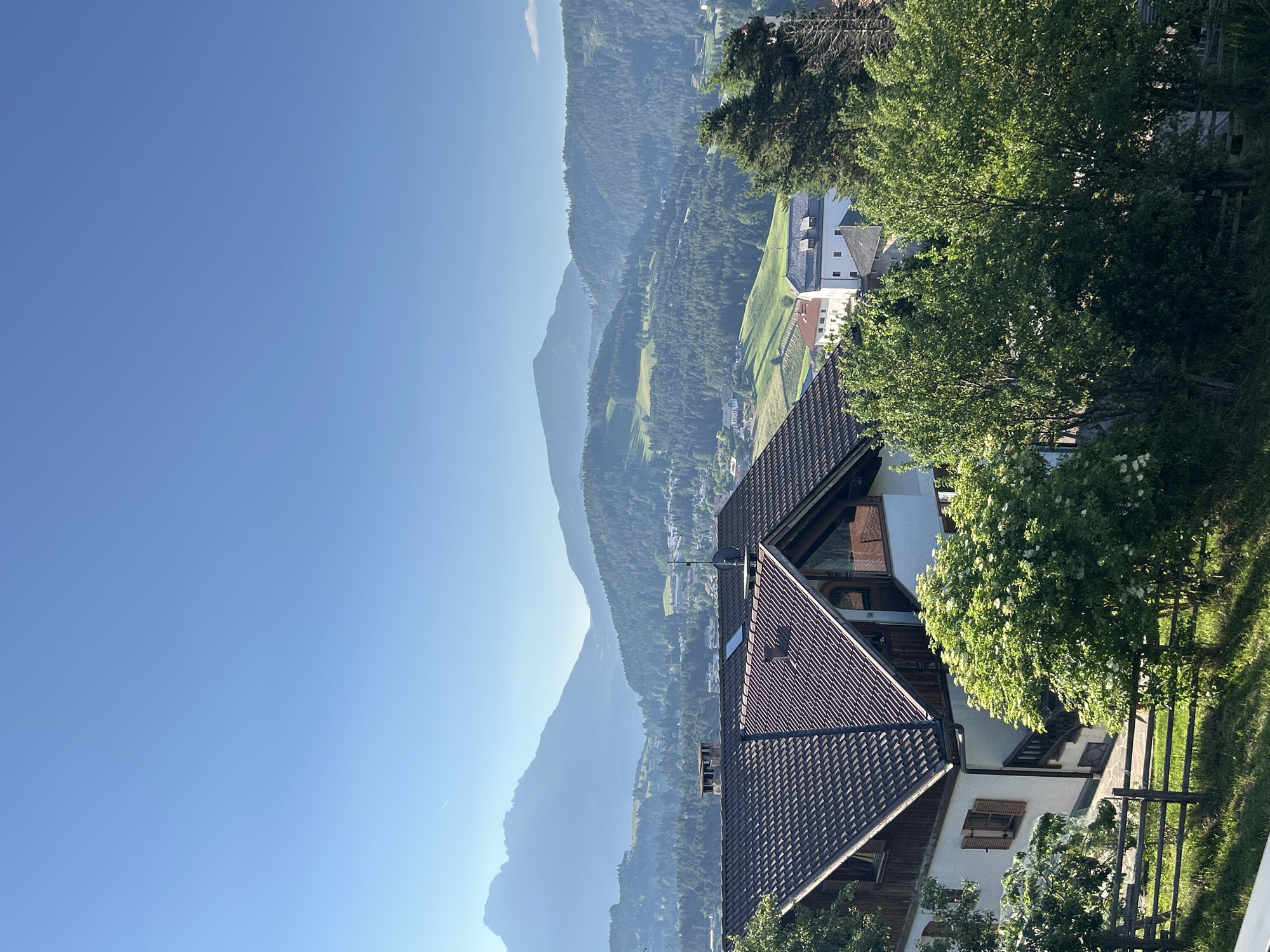 The image depicts a picturesque mountain landscape under a clear blue sky. In the foreground, a traditional house with a dark tiled roof is visible, featuring wooden shutters on its windows. Adjacent to the house, a wooden fence encloses a grassy area, and several lush green trees are in bloom. Beyond the house, the landscape unfolds into rolling hills covered in dense green forests, interspersed with scattered buildings and fields. In the distance, majestic mountains are silhouetted against the hazy horizon, their peaks softened by atmospheric perspective. The overall scene evokes a sense of tranquility and natural beauty, typical of a rural mountain village.