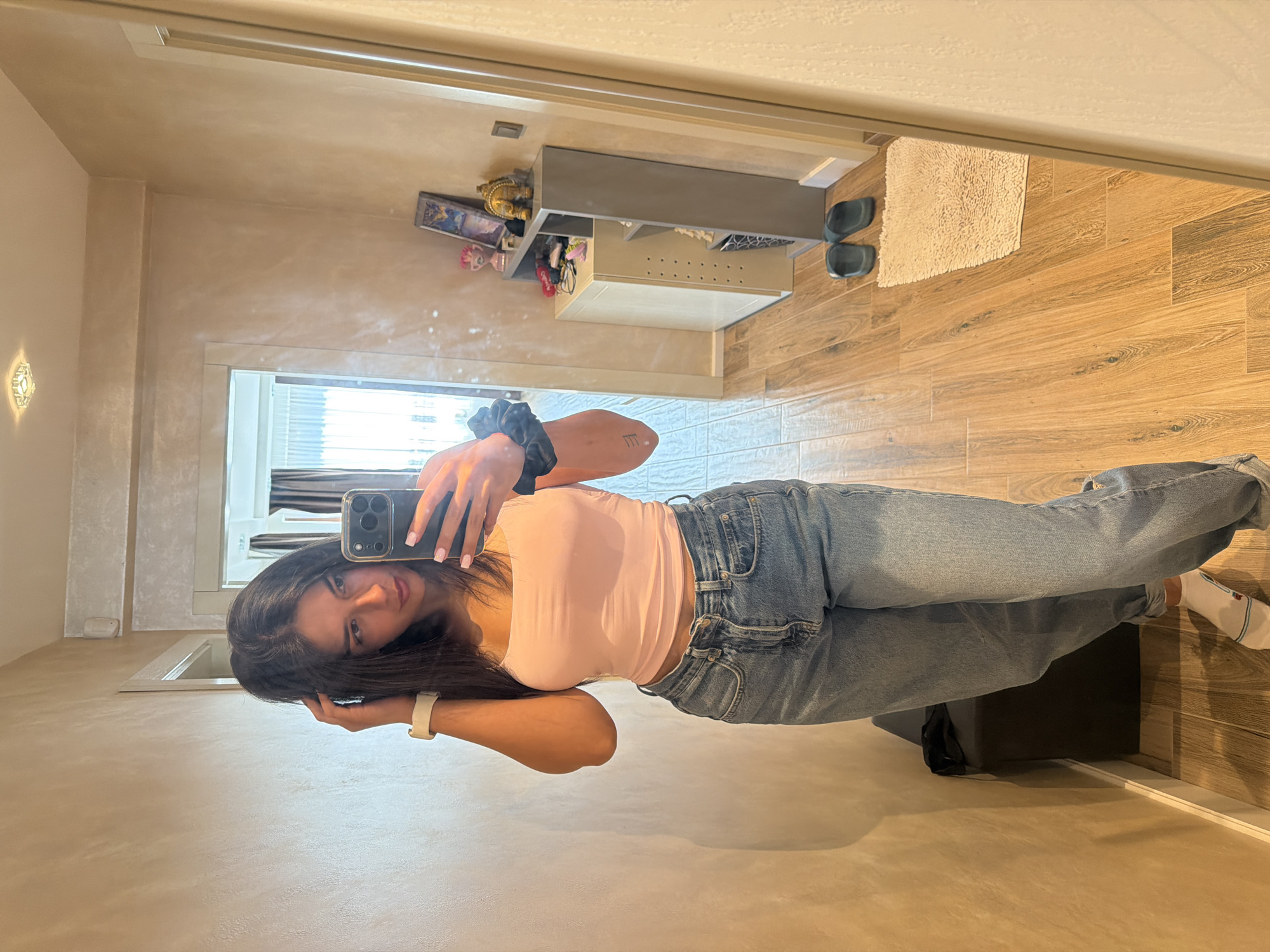 A young woman stands in a hallway and takes a mirror selfie. She has dark hair and is wearing a pink tank top and blue jeans. She is holding her phone in her right hand and touching her hair with her left hand. The hallway is lit by overhead lights and has wooden floors. There is a mirror on the right side of the image, and a doorway leading to another room on the left. There is a small shelf with decorative items and a pair of dark blue slides on the floor. The scene appears to be indoors, likely in a residential setting. There is no information to indicate this location is in the city of Gabrovo, Bulgaria.