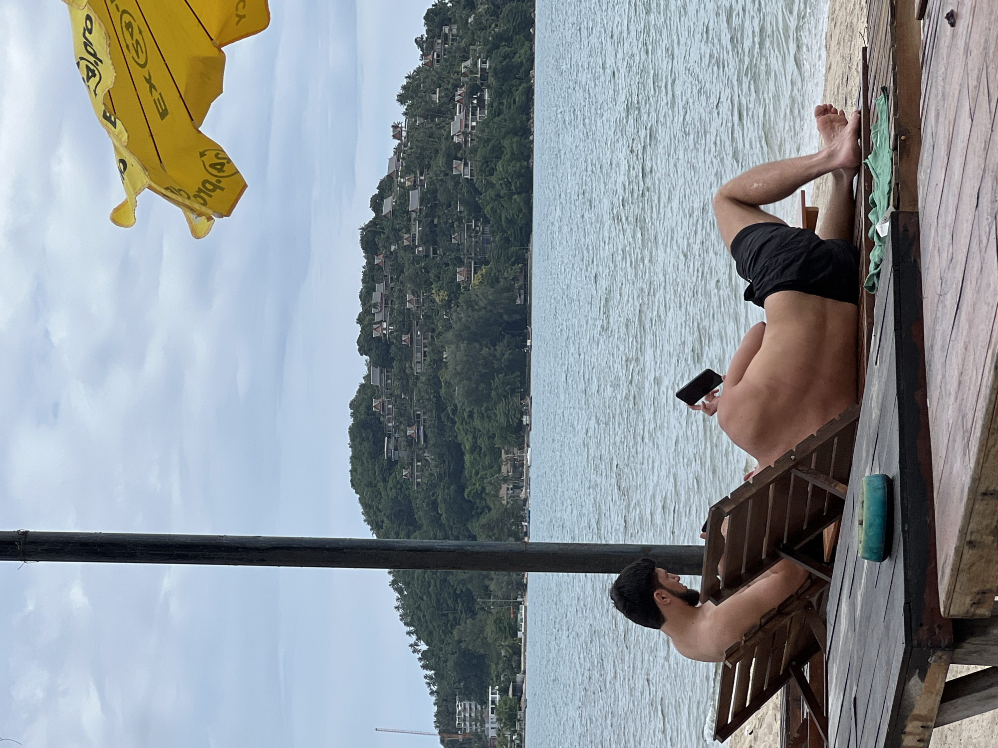 In an outdoor scene with a cloudy sky, two shirtless men are relaxing on lounge chairs on a beach. The man on the left is facing away from the camera, looking down at a black object, possibly a phone, in his lap. He has dark hair and a beard. The man on the right is lying on his back, facing away from the camera, holding a black phone in his right hand. He is wearing black shorts. In the foreground, a weathered wooden table is visible with a blue ashtray on it. Behind the men, the ocean stretches out to a lush, green hillside dotted with buildings. A yellow umbrella is partially visible in the upper right corner of the frame.