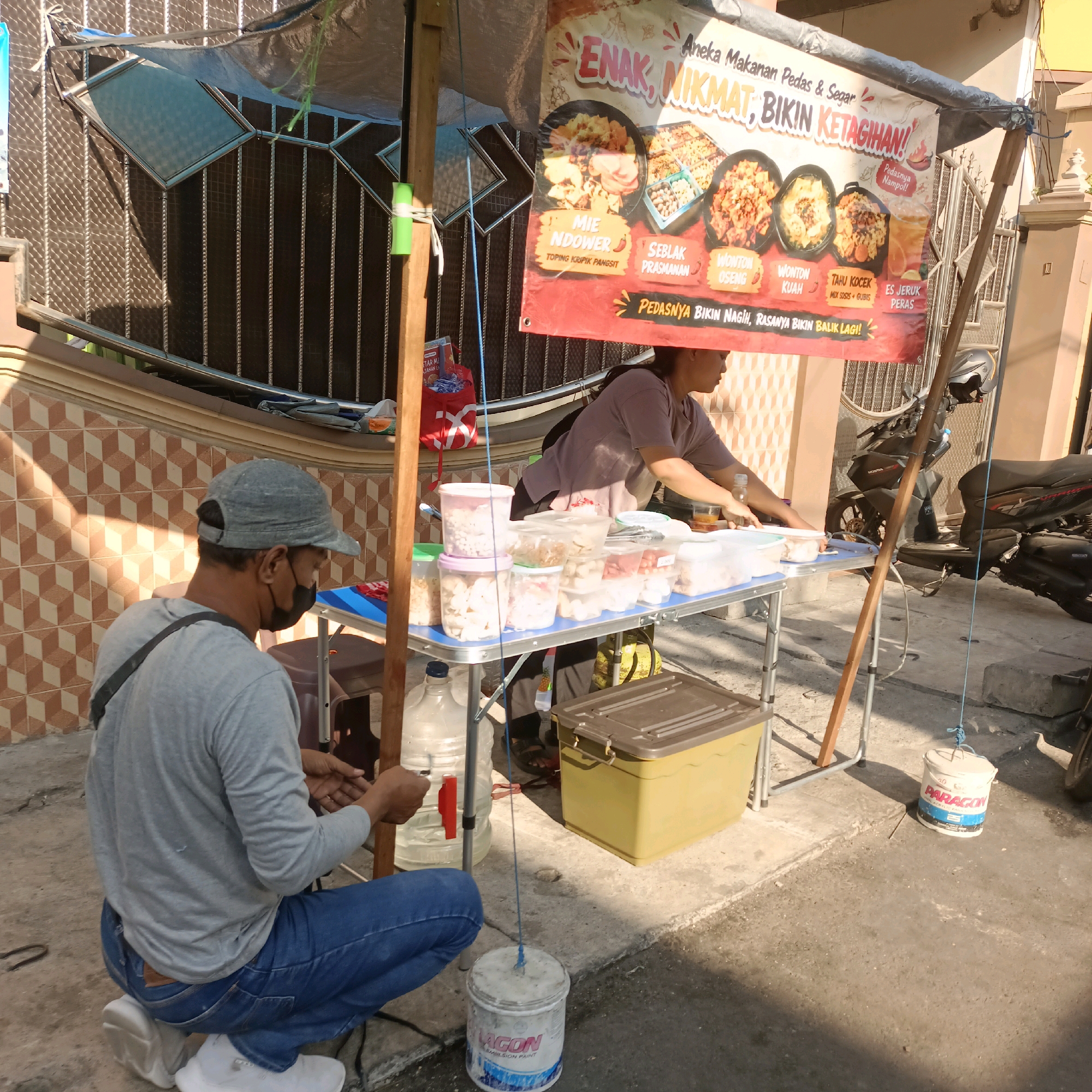 An outdoor food stall operates under a makeshift canopy in Surabaya, Indonesia. A man wearing a grey t-shirt, blue jeans, and a grey baseball cap is kneeling on the ground, facing away from the camera, with a black face mask pulled down below his chin. He appears to be tying something with a blue string. A woman, wearing a purple t-shirt, is standing behind a blue table, interacting with food containers. The table is laden with various plastic containers filled with food items and drinks. A large banner displaying food images and text in Indonesian hangs above the stall. In the background, a motorcycle is parked, and the facade of a building with tiled walls is visible. The ground is paved with concrete. A white bucket with the brand name "PARAGON" is placed on the ground.