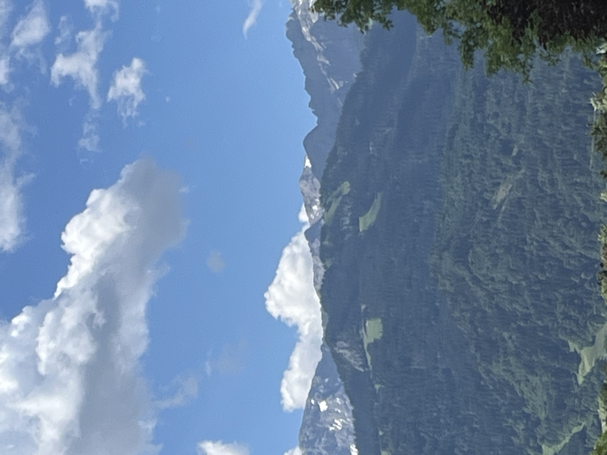 A distant shot captures the majestic Alps mountains in Garmisch-Partenkirchen, Germany, on a bright, sunny day. The sky is a vibrant blue, dotted with fluffy white clouds. The rugged mountain peaks are partially covered with snow, contrasting with the lush green slopes below. A dense forest blankets the lower parts of the mountains, with patches of lighter green indicating cleared areas or meadows. The scene is serene and picturesque, conveying the natural beauty of the Bavarian Alps.