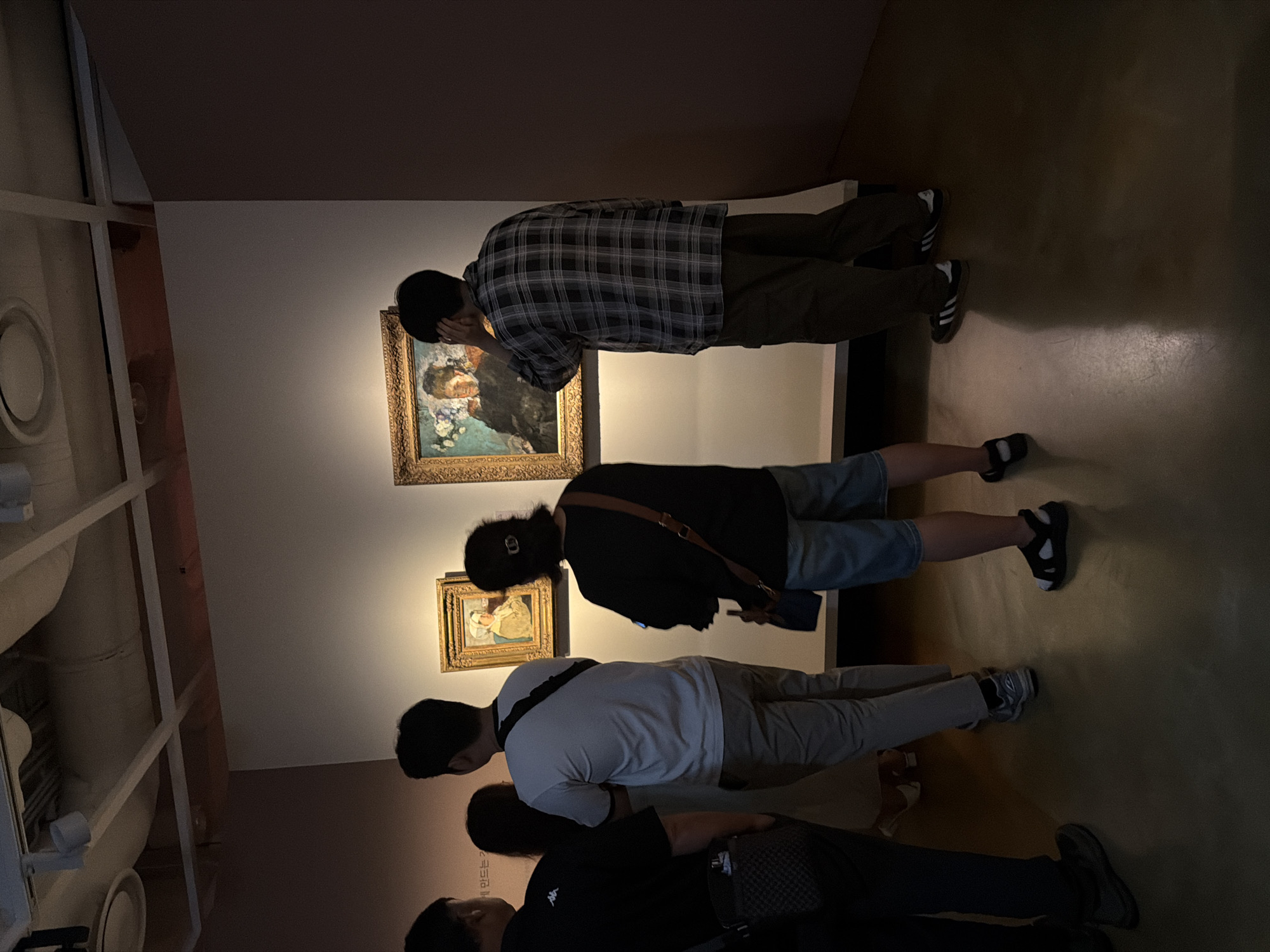 In the dimly lit interior of an art gallery, likely located in Seoul, South Korea, four individuals are observed examining paintings displayed on a light-colored wall. On the left, a person wearing a black t-shirt with a logo is partially visible from the back, holding a bag. Next to them, a woman in a light-colored dress stands, her back to the viewer. To her right, a man in a white t-shirt and khaki pants is facing the paintings, with a black strap across his chest. On the far right, another man, wearing a plaid shirt and dark pants, is seen from behind, one hand touching his ear as he looks at a painting. Two framed artworks are visible on the wall: a smaller painting to the left and a larger one to the right, both illuminated by spotlights from above. The floor is a dark, polished surface reflecting the ambient light. The ceiling features exposed ductwork and lighting fixtures.
