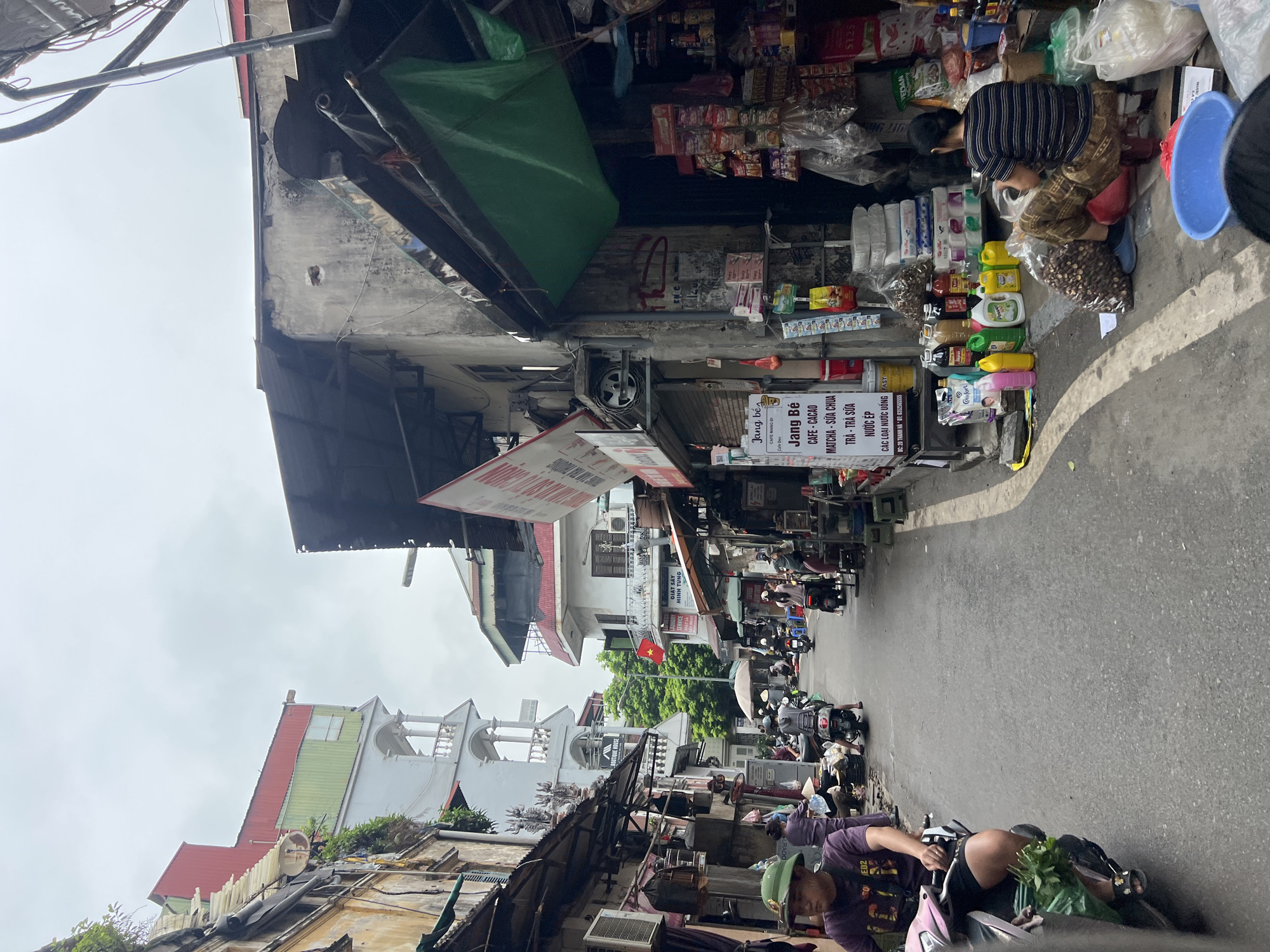 The image captures a street scene in Hanoi, Vietnam, likely in an older, more densely populated district. The perspective is from street level, looking down a narrow lane flanked by buildings of varying architectural styles and conditions. The buildings on either side are packed closely together, with multiple stories and balconies, some appearing aged with peeling paint and exposed brickwork, while others are newer and more brightly colored. Several shops and market stalls line the street, displaying a variety of goods, including food items, drinks, and household products.
The street itself is paved and appears to be a busy thoroughfare for motorbikes and pedestrians. Several motorbikes are visible, with some people riding them and others parked. A man in a purple t-shirt and a green hat is seated on a pink motorbike in the foreground, with a bunch of leafy greens in a plastic bag on the front of his bike. Further down the street, other individuals are seen riding motorbikes or walking amongst the stalls.
A prominent sign, written in Vietnamese, is visible on a building on the left side of the street, indicating a cafe named "Jang Bé" which offers items like "Cafe Cacao," "Matcha - Sua Chua," "Tra - Tra Sua," and "Nuoc Ep." A Vietnamese flag is also visible, fluttering from a pole on one of the buildings.
The overall atmosphere suggests a vibrant, bustling urban environment typical of many Vietnamese cities, characterized by its narrow streets, diverse architecture, and active street life. The weather appears to be overcast, with a cloudy sky.