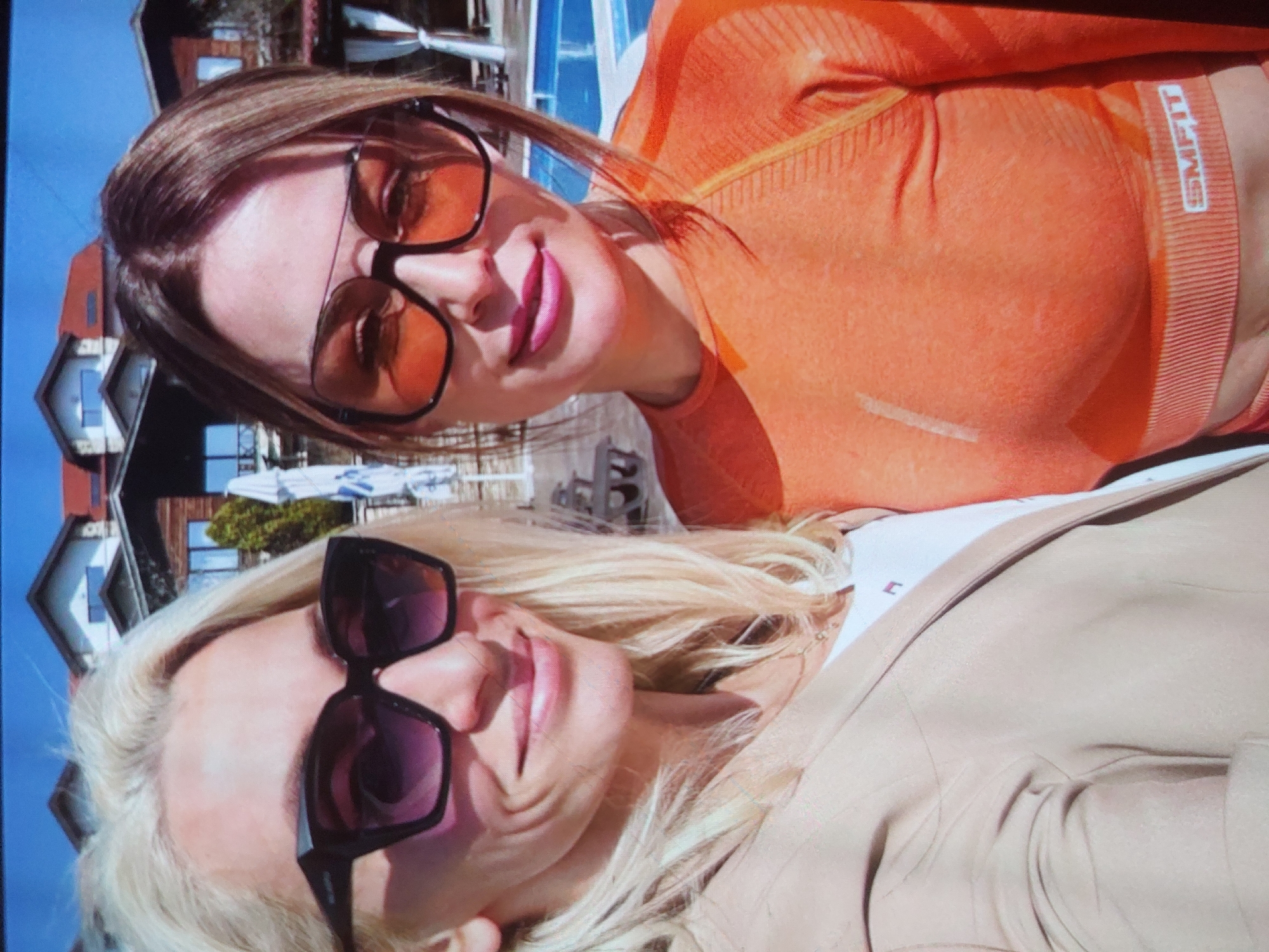 Two smiling women pose for a close-up outdoor photograph, likely a selfie, on a bright, sunny day in Tsarevo, Bulgaria. The image is rotated 90 degrees counter-clockwise.
In the foreground, a blonde woman with loose hair wears large dark-framed sunglasses with dark lenses. She is dressed in a light-colored, possibly beige or khaki, jacket over a white top. Her face shows a broad, happy smile.
Behind her, slightly out of focus and positioned higher in the frame, is a woman with reddish-brown hair. She wears large dark-framed sunglasses with orange-tinted lenses and bright pink lipstick. She is wearing a fitted, long-sleeved orange athletic top. On the elastic band of her top, the brand name "SMFIT" is clearly visible in white letters. She also has a gentle smile.
The background, largely out of focus, reveals elements of a coastal or resort setting, consistent with Tsarevo. Several buildings with pitched roofs are visible, along with some distant greenery. The blue color dominating the top left of the original image suggests either a clear blue sky or a body of water, such as the Black Sea, typical for this Bulgarian location. The overall mood is cheerful and relaxed, reflecting a pleasant outdoor experience.