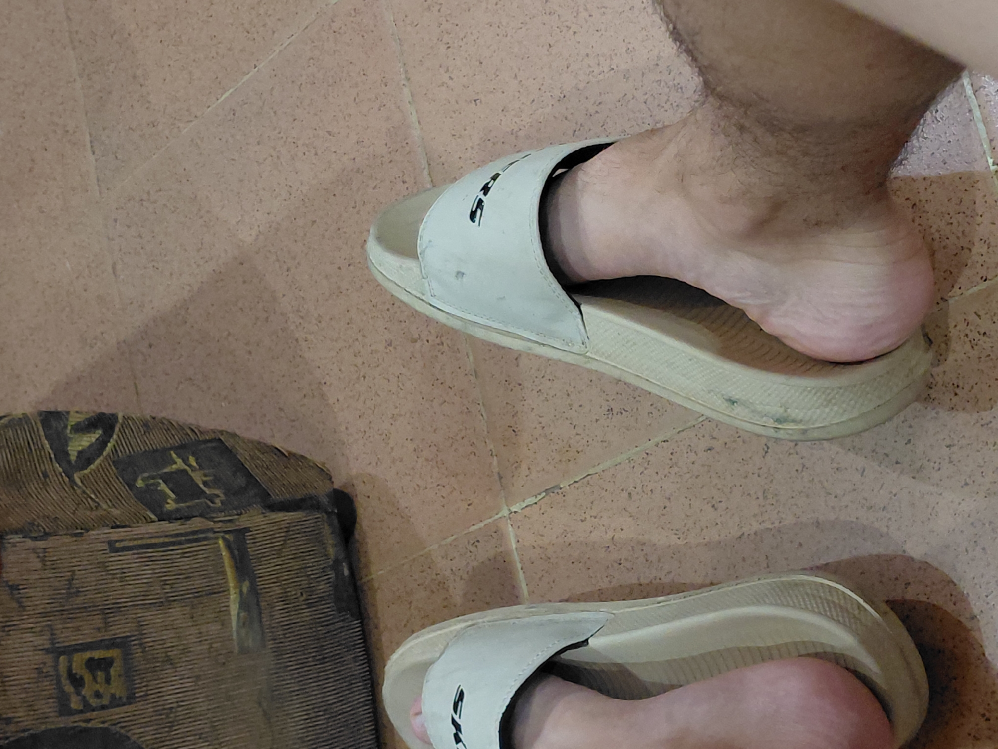 An overhead shot depicts a person's feet, wearing light-colored slide sandals, resting on a tiled floor. The floor is composed of reddish-brown, speckled square tiles with visible grout lines that show some wear. The upper foot is partially inserted into a sandal, with the heel slightly raised, revealing the foot's arch and some body hair on the lower leg/ankle area. The strap of this sandal displays partial dark text, appearing to be "SKE", indicative of the brand Skechers. Both sandals are light beige or off-white and show signs of use and minor dirt. The second foot is mostly obscured but also wears a similar sandal. In the bottom left corner, a dark, textured object, possibly a woven mat or bag, with abstract patterns in lighter hues (yellow, black, gray) is visible. The scene is indoors, likely in a domestic setting, located in Santa Ana, Venezuela. No specific activity or emotion is discernible.