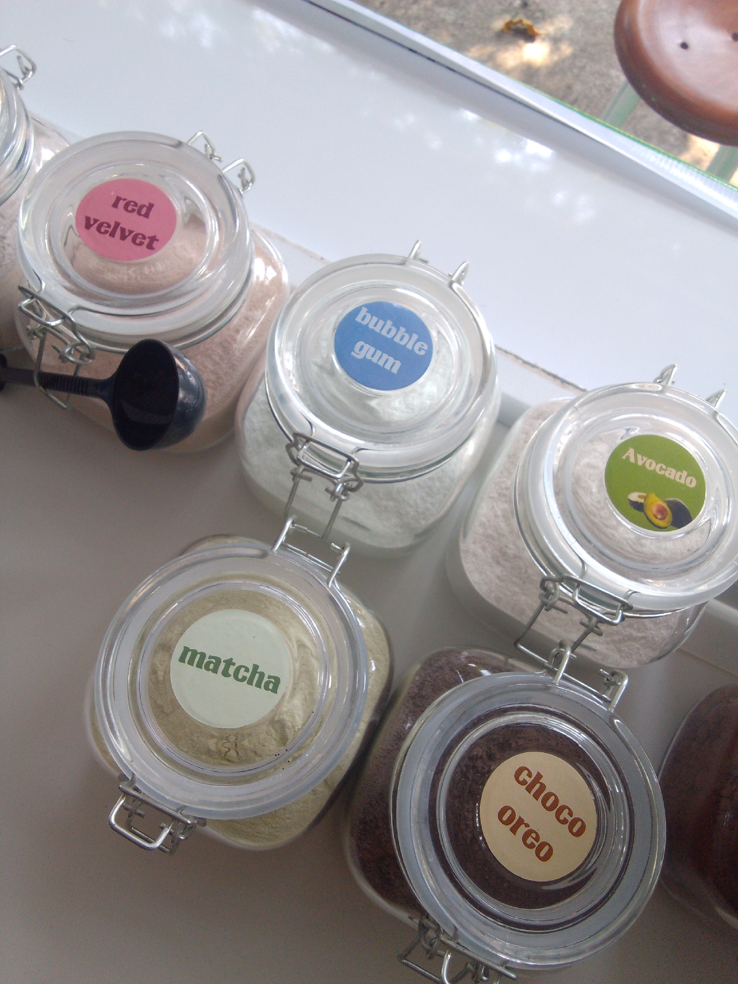 This is an eye-level, medium shot of several glass jars filled with powdered substances, arranged on a white surface near a window. Each jar has a metal clasp closure and a circular label on the lid indicating the contents. From left to right, the visible labels read "red velvet", "bubble gum", "Avocado", "matcha", and "choco oreo". A black plastic scoop is resting in the "red velvet" jar. The background shows a blurred outdoor scene with greenery and a brown object, possibly a piece of furniture. The lighting suggests an indoor setting with natural light coming from the window. There is no indication of the specific location in Trenggalek, Indonesia.