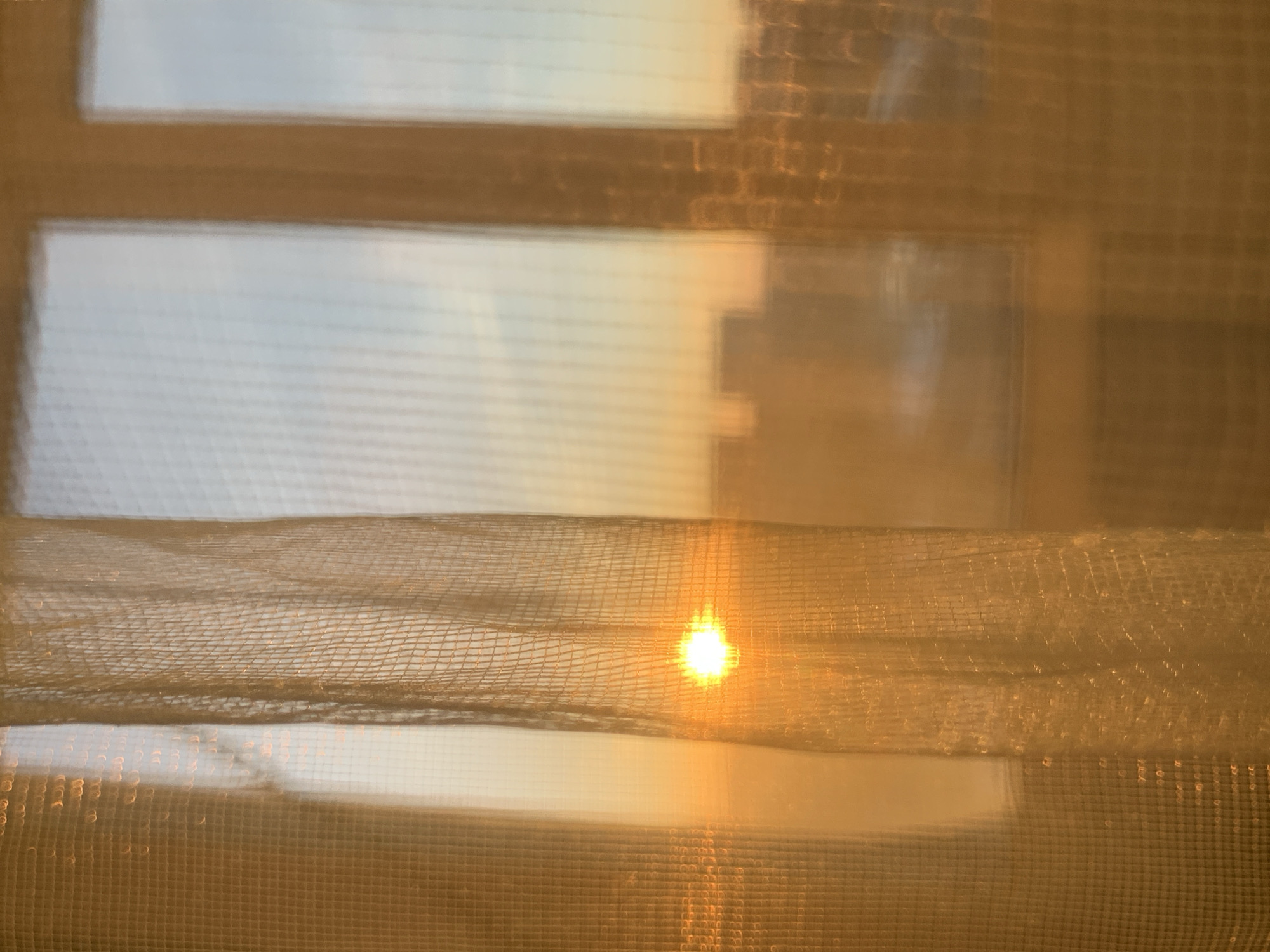 The image shows a window with sheer curtains. Sunlight is streaming through the window, creating a bright spot on the curtain. The view outside the window is blurry, but suggests buildings or other structures. There is no information in the image to identify this location as the city of Izhevsk, Russia. There is no information in the image about people, subjects, actions, or interactions.