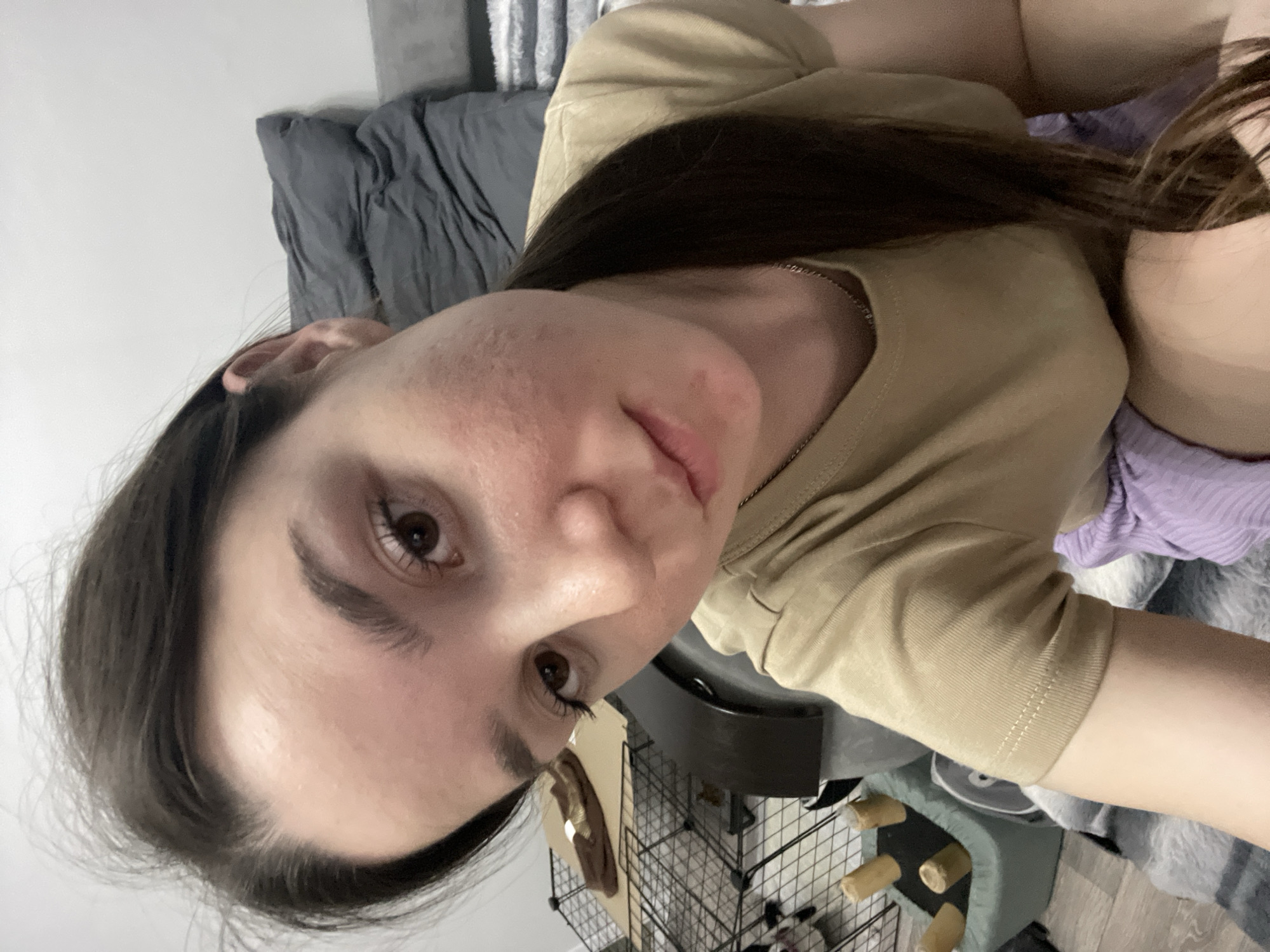 A woman with light skin and dark brown hair is lying on a bed with gray sheets, wearing a beige t-shirt and a silver necklace. Her face is close to the camera, with visible pores and slight redness on her cheeks. Her eyes are brown, and she appears to be looking directly at the camera. Behind her, there is a wire cage with objects inside, possibly a pet cage. The overall impression is that she is relaxed and casual. There is no indication of location in Chaikovsky, Russia.