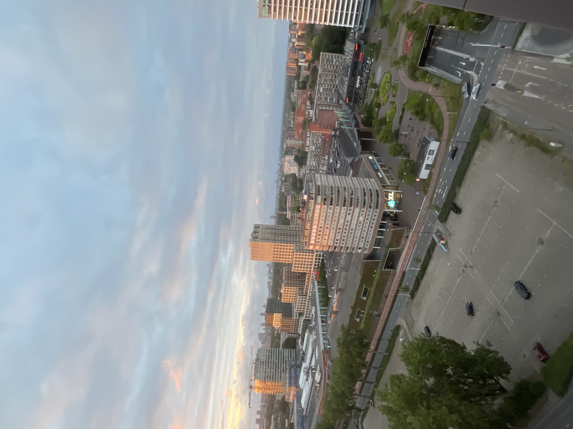 An aerial shot captures a city scene at dusk, likely in Europe, given the architecture. The sky is a gradient of pale blue and grey clouds, with hints of pink and orange from the setting sun on the left side. In the foreground, a partially visible tall building with white horizontal siding stands on the right. Below, a road with traffic lights and vehicles leads into a tunnel entrance on the right. A parking lot is visible to the left of the road, with several cars parked and moving. Further into the scene, a row of trees lines a street. Beyond the trees, various buildings of different heights and architectural styles are present, including a prominent building with a curved facade in the mid-ground, reflecting the sunset. In the background, a sprawling cityscape extends to the horizon. The overall atmosphere is serene, with the soft lighting of dusk casting long shadows and highlighting the urban landscape.
