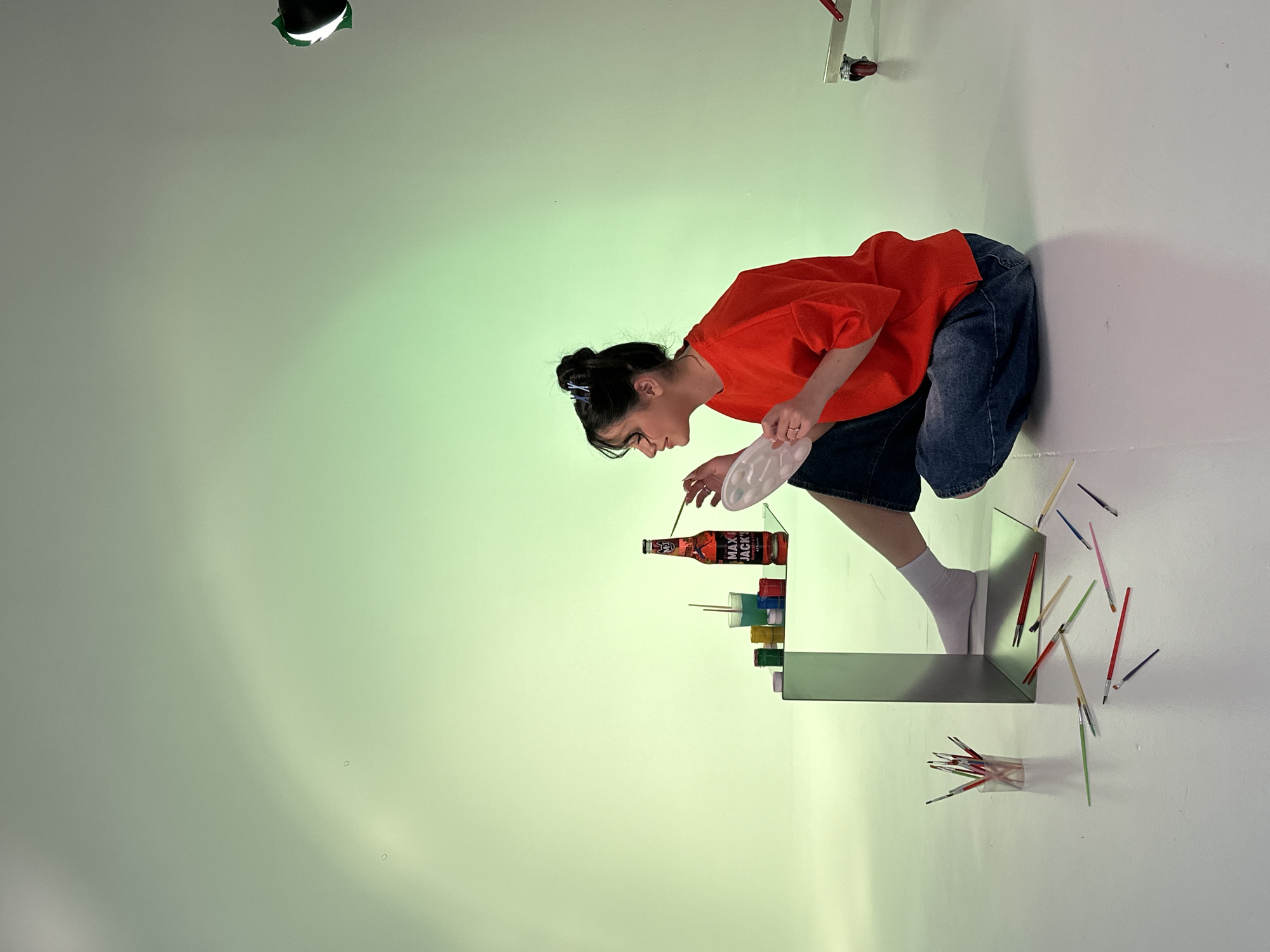 A female with dark hair, wearing an orange shirt and jeans, is standing on a shelf. She is holding a palette in her left hand and a paintbrush in her right, appearing to paint on a bottle of "MAX JACK'S". The bottle and several stacked paint jars are also on the shelf. To the right of the shelf are scattered paintbrushes. The background is a gradient of green and white. A spotlight is visible in the upper left corner of the image. The location is not evident from the image; it is not possible to verify that it is in Yerevan, Armenia.
