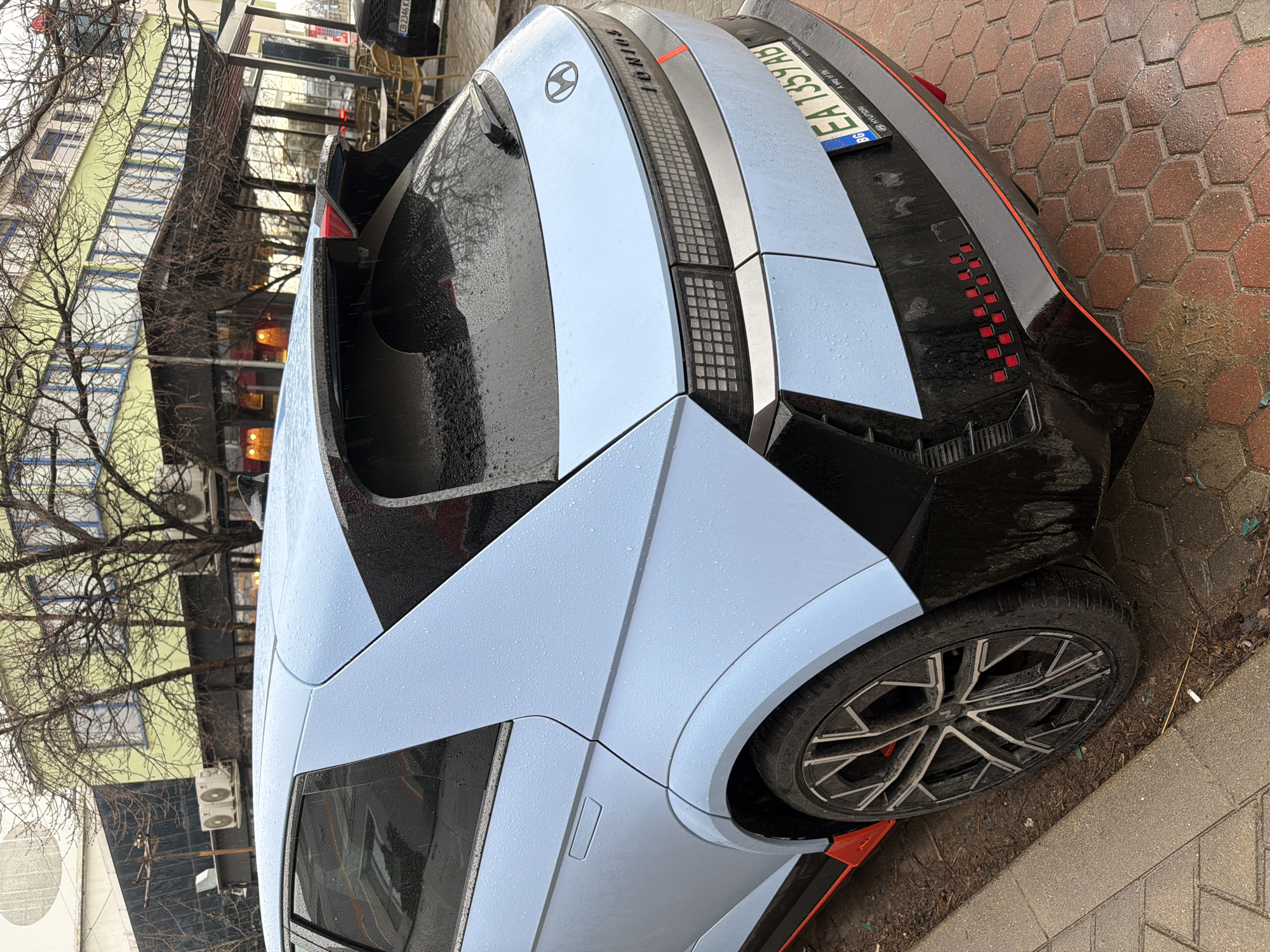 The image shows the rear of a light blue Hyundai Ioniq 5 N car parked on a brick-paved sidewalk in Sofia, Bulgaria. The car's license plate reads "EA 1359 AB". The background features a building with green and white walls and bare trees. The weather appears to be damp, with water droplets visible on the car's surface. The car's black spoiler and distinctive rear light design are visible. The wheel has an intricate multi-spoke pattern.