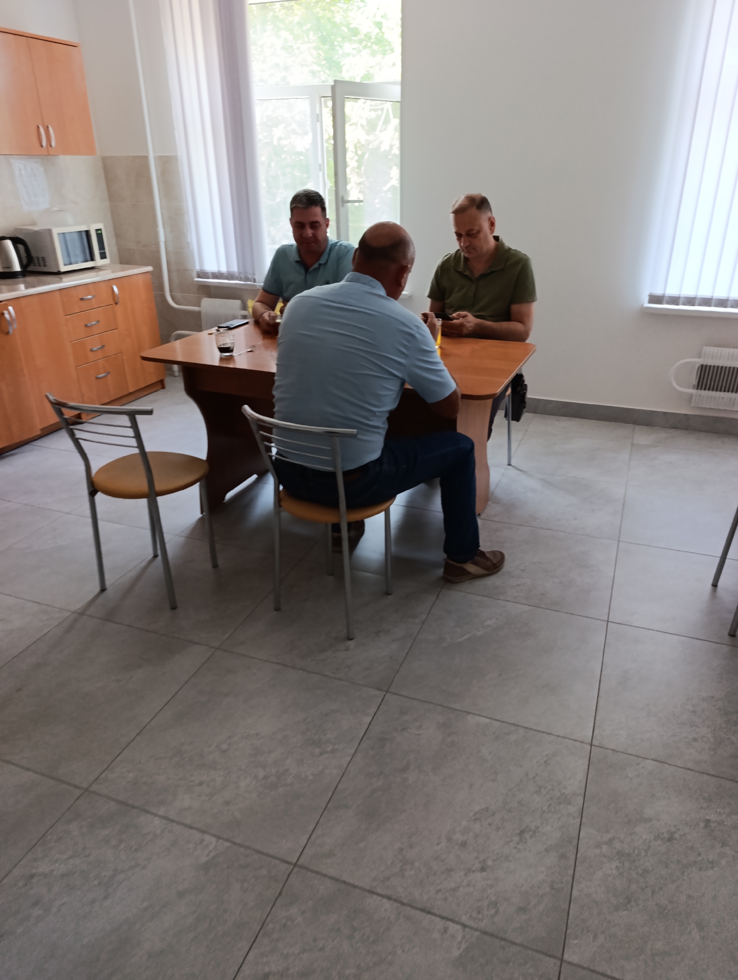 In a room resembling a break room or office common area, three men are gathered around a wooden table. The room features a kitchenette on the left with light wood cabinets, a microwave, and a kettle. The floor is tiled with large grey tiles. Natural light streams in from windows on the right side of the image. The men are seated, and one appears to be using a mobile phone. The overall scene depicts a casual gathering in a functional, somewhat dated, interior. This scene is located in the town council of Dubăsari, Moldova.