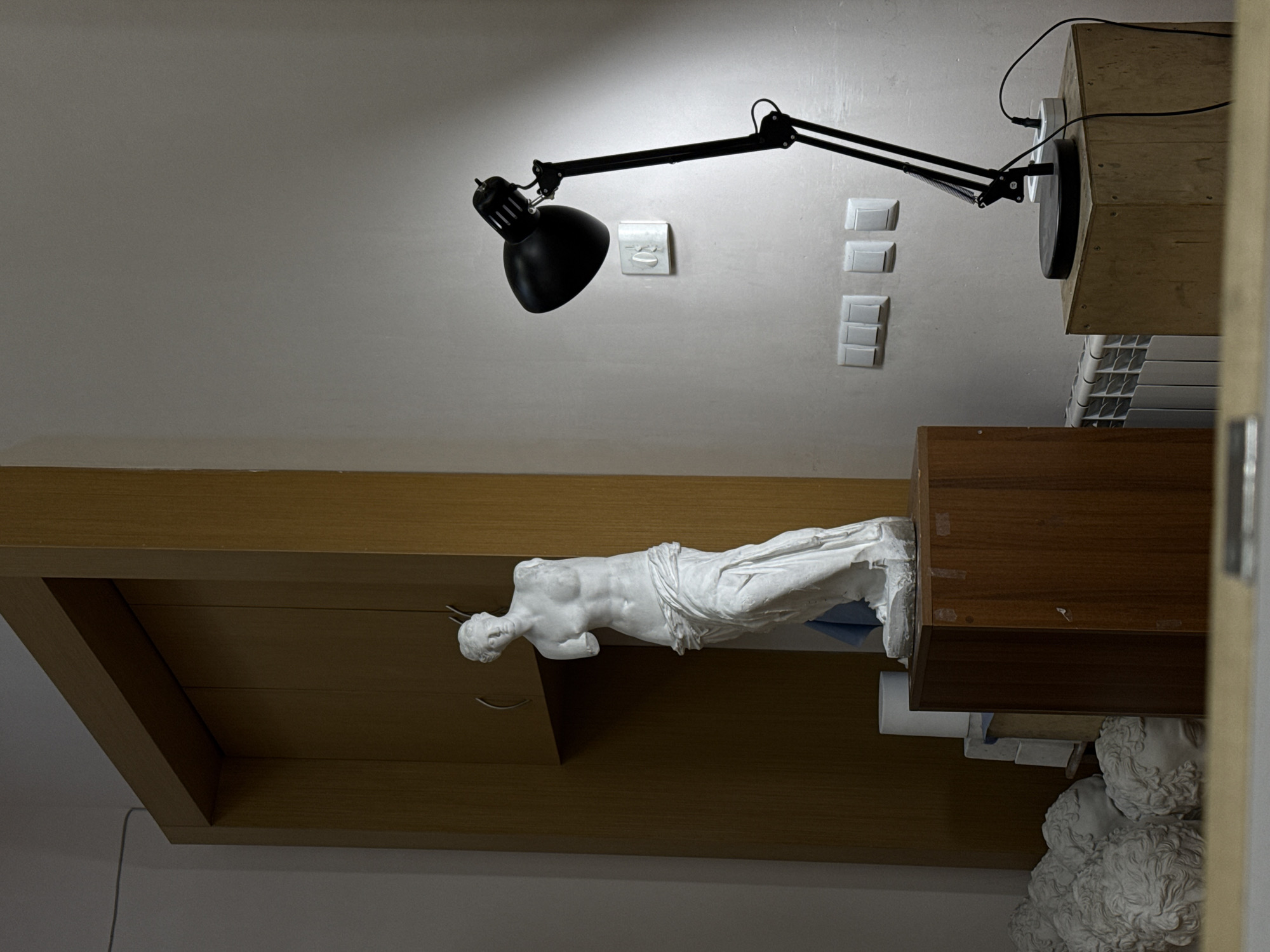 The image shows an interior scene. In the upper portion of the frame is a white wall with a black adjustable desk lamp attached to it. Adjacent to the lamp are a light switch and a power outlet. Below the lamp, a wooden shelf is mounted, on which lies a white statue of a reclining nude female figure. The bottom of the frame shows parts of other statues. The image is rotated sideways. There is no verifiable evidence to determine the location as Yerevan, Armenia.