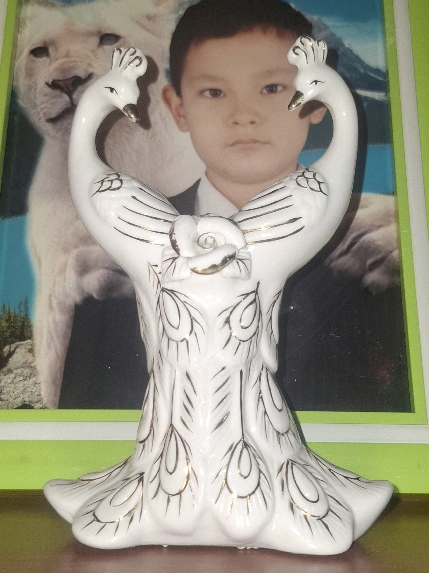 A white ceramic figurine of two peacocks with gold accents stands on a wooden surface. The figurine is in the foreground and is centrally located. Behind it, a framed picture is visible. The picture features a white lion on the left, and a young boy in a suit on the right. The background of the picture appears to be a landscape with mountains and a body of water. The frame of the picture is bright green. The overall scene is indoors and appears to be a domestic setting.