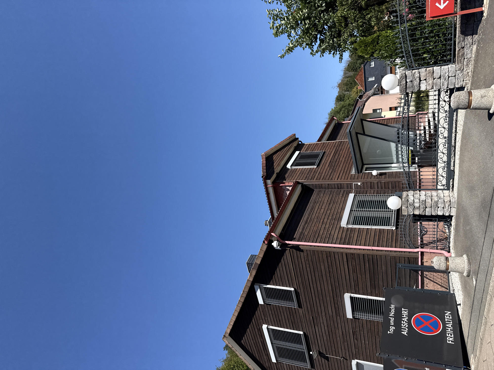 A low-angle outdoor shot captures a clear, bright blue sky above a brown wooden house in Bad Lauterberg im Harz, Germany. The house features dark brown horizontal siding, white-trimmed windows with horizontal blinds, and a red roof with visible gutters. A pink pipe runs vertically along the side of the house. To the left of the house, a black sign displays German text: "Tag und Nacht AUSFAHRT" (Day and Night EXIT) and "FREIHALTEN" (KEEP CLEAR) with a red and blue circular "no stopping" symbol. A black metal fence with decorative scrollwork borders the property in front of the house, leading to a paved walkway. Stone pillars topped with white spherical lights mark the entrance. Trees are visible in the background on a hillside.