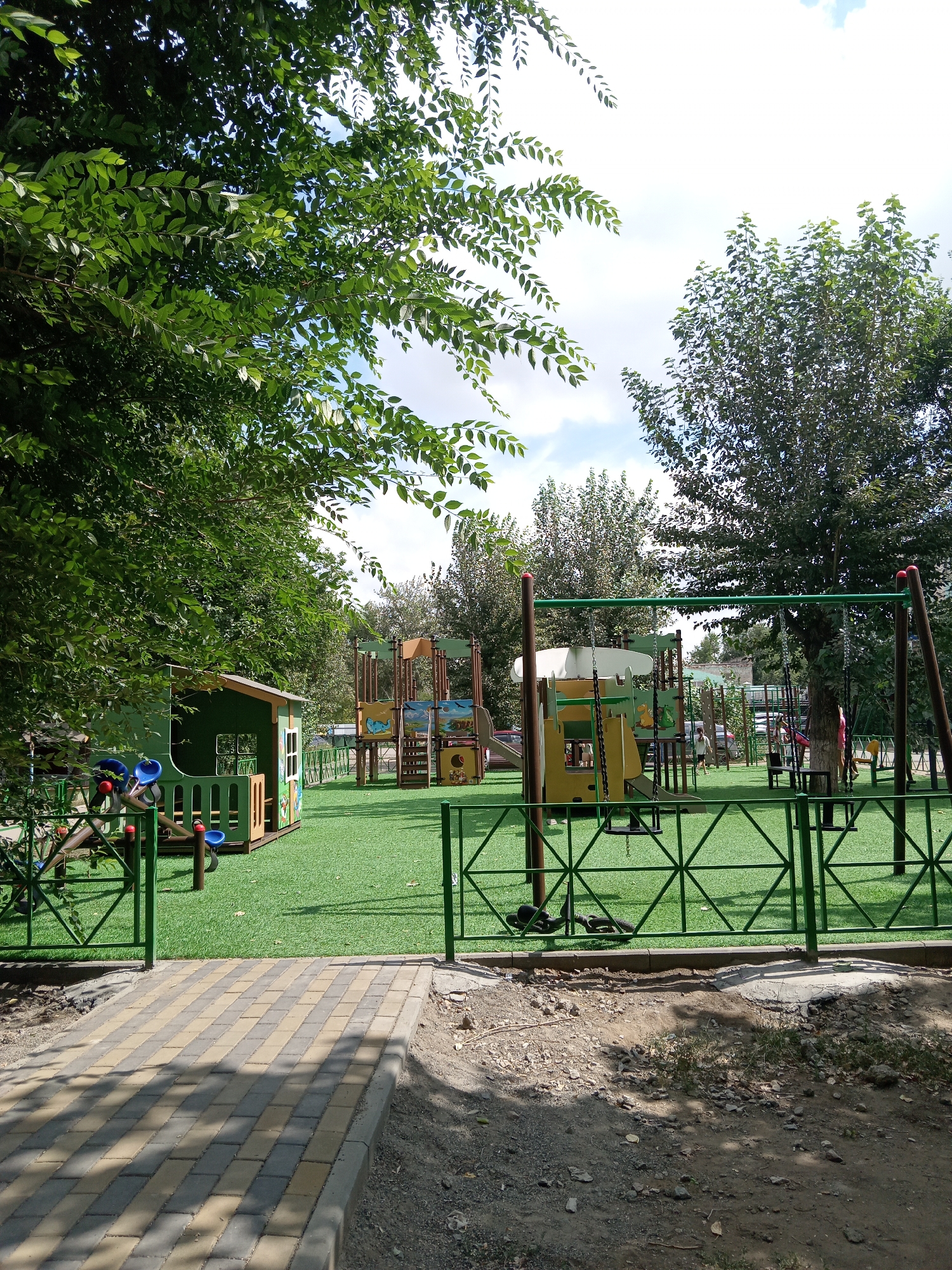 The image depicts an outdoor children's playground located in the city of Rudny, Kazakhstan. The playground features artificial turf flooring and a green metal fence enclosing the play area. Several play structures are visible, including a small green playhouse with blue slides and a larger multi-colored play structure with slides, swings, and climbing elements. Trees with green foliage surround the playground, indicating a summer or late spring setting. A paved pathway leads to the entrance of the playground. The sky is partially cloudy. Two children are present in the play area, one in a pink shirt and the other in a black shirt.