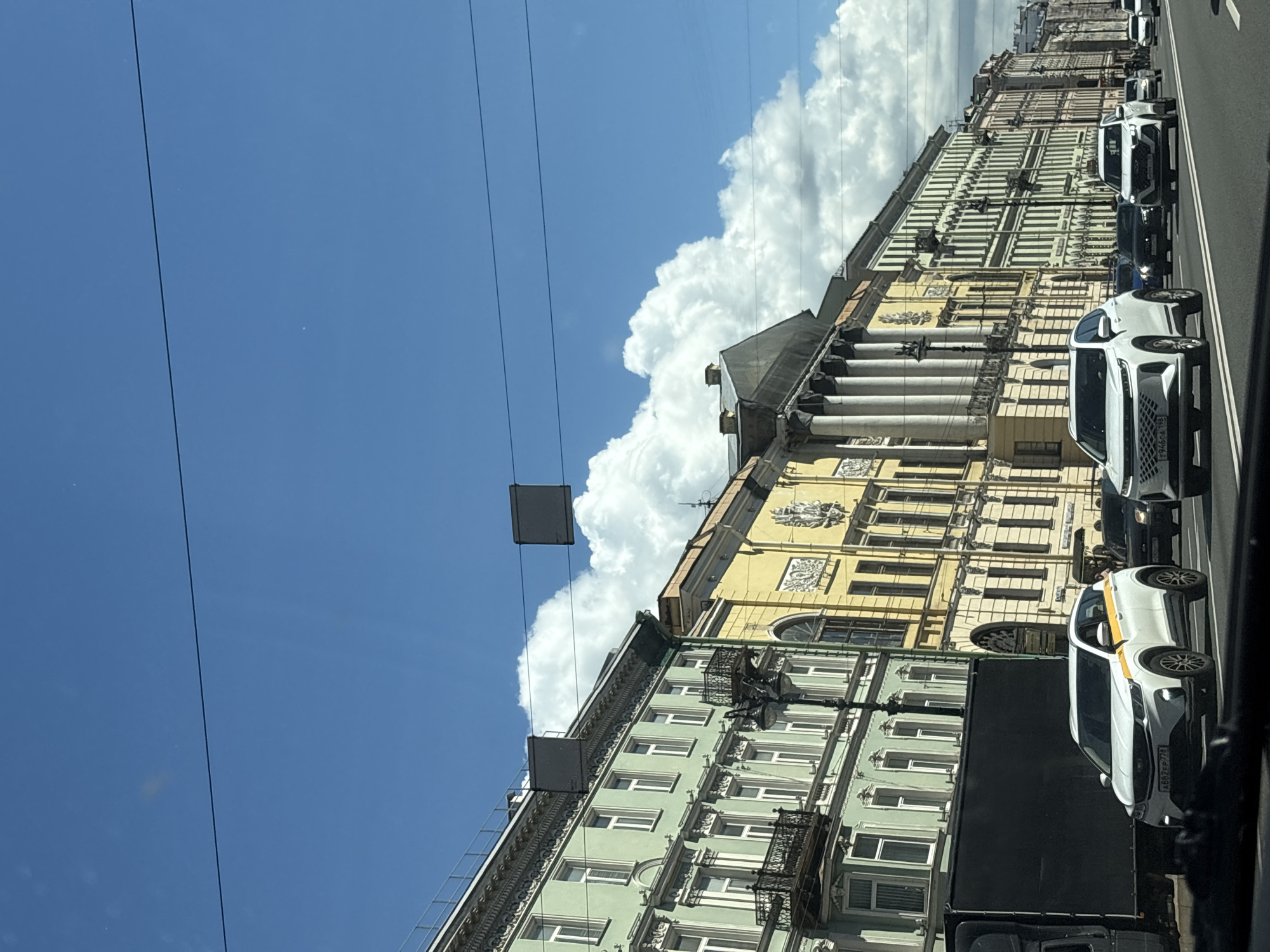 The image depicts a street scene in Saint Petersburg, Russia, likely during daylight hours on a clear day with scattered clouds. The perspective is from inside a vehicle, looking forward.
On the right side of the street, a row of ornate, multi-story buildings with distinct architectural styles lines the road. The buildings feature a variety of colors, including light green, yellow, and pastel shades, with detailed facades, large windows, and decorative elements. One building, in particular, has a prominent portico with classical columns.
The street itself is paved and appears to be a multi-lane thoroughfare. Several cars are visible, driving in the same direction as the viewer. These include a white SUV with yellow accents on the side, a white modern SUV, and darker vehicles.
In the sky, a bright blue sky is visible with large, fluffy white clouds. Overhead, there are dark power or communication lines stretched across the frame, with two dark rectangular objects suspended from them.
On the left side of the image, part of a large, dark-colored truck or trailer is visible, partially obscuring the buildings.
The overall impression is of a busy urban environment with a mix of historical architecture and modern traffic under a bright sky.