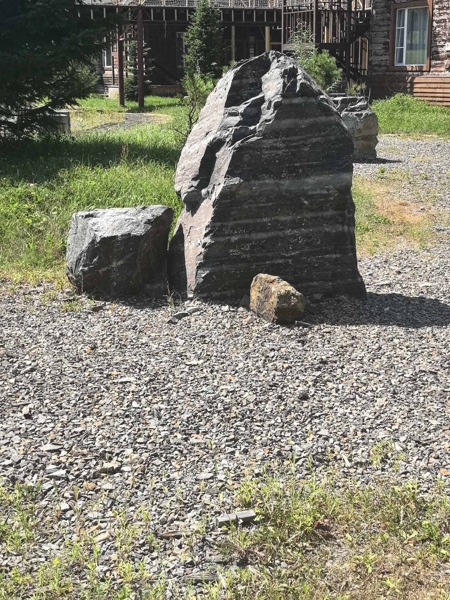 In Solnechnyi, Russia, a scene features several large rocks arranged on a gravel surface. The primary subject is a tall, dark gray boulder with distinct horizontal striations, standing upright. To its left, a smaller, lighter gray boulder is positioned. In front of the main boulder, a single, smaller, reddish-brown rock rests on the gravel. The background shows a grassy area with some vegetation, and a wooden building with a dark-colored roof and siding is visible behind the rocks. The overall environment appears to be an outdoor setting, possibly a park or a landscaped area, under daylight conditions.