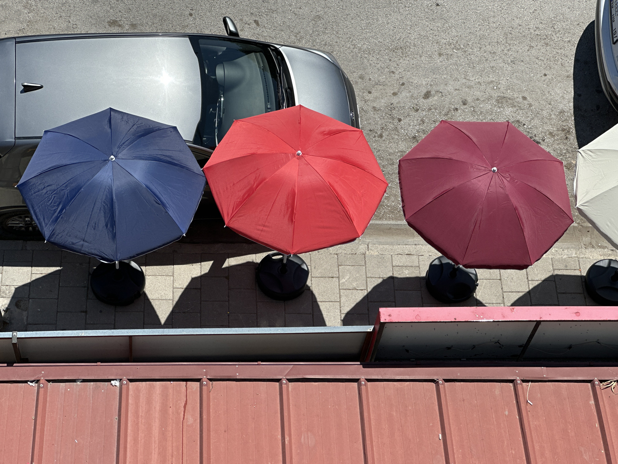 An aerial shot reveals three open umbrellas casting shadows, positioned next to a silver car and adjacent to a weathered, corrugated roof. The umbrellas are blue, red, and burgundy. All items are positioned on a paved surface, presumably the exterior of a building in Fethiye, Turkey.