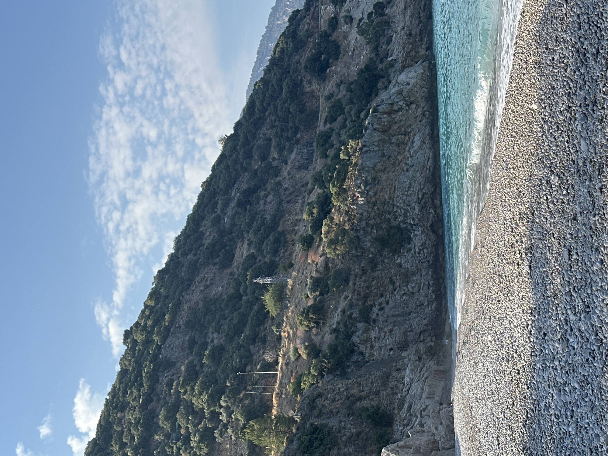 The image depicts a serene, sun-drenched beach with a mountainous backdrop, likely located in Fethiye, Turkey. The foreground is dominated by a vast expanse of smooth, grey pebbles that form the beach. Gentle waves, tinged with turquoise hues, lap at the shore, leaving a thin, foamy white line where they meet the pebbles.
The coastline curves gently, leading the eye towards a rocky cliff face that rises steeply from the water's edge. This cliff is rugged and textured, with patches of dry grass and scattered green vegetation clinging to its slopes.
Further up the mountainside, a dense cover of dark green trees, possibly olive or pine trees, blankets the terrain. The mountain itself is steep and imposing, with its summit disappearing into the clear blue sky. Faint clouds, wispy and white, are scattered across the sky, suggesting a pleasant day.
On the mid-slope of the mountain, several man-made structures are visible, including two tall, slender poles that could be utility poles or lighting fixtures, and a more complex metal structure resembling a transmission tower. A winding path or road is also discernible etched into the mountainside. The overall scene conveys a sense of natural beauty and tranquility, typical of the coastal regions of Fethiye. No people are visible in the image.