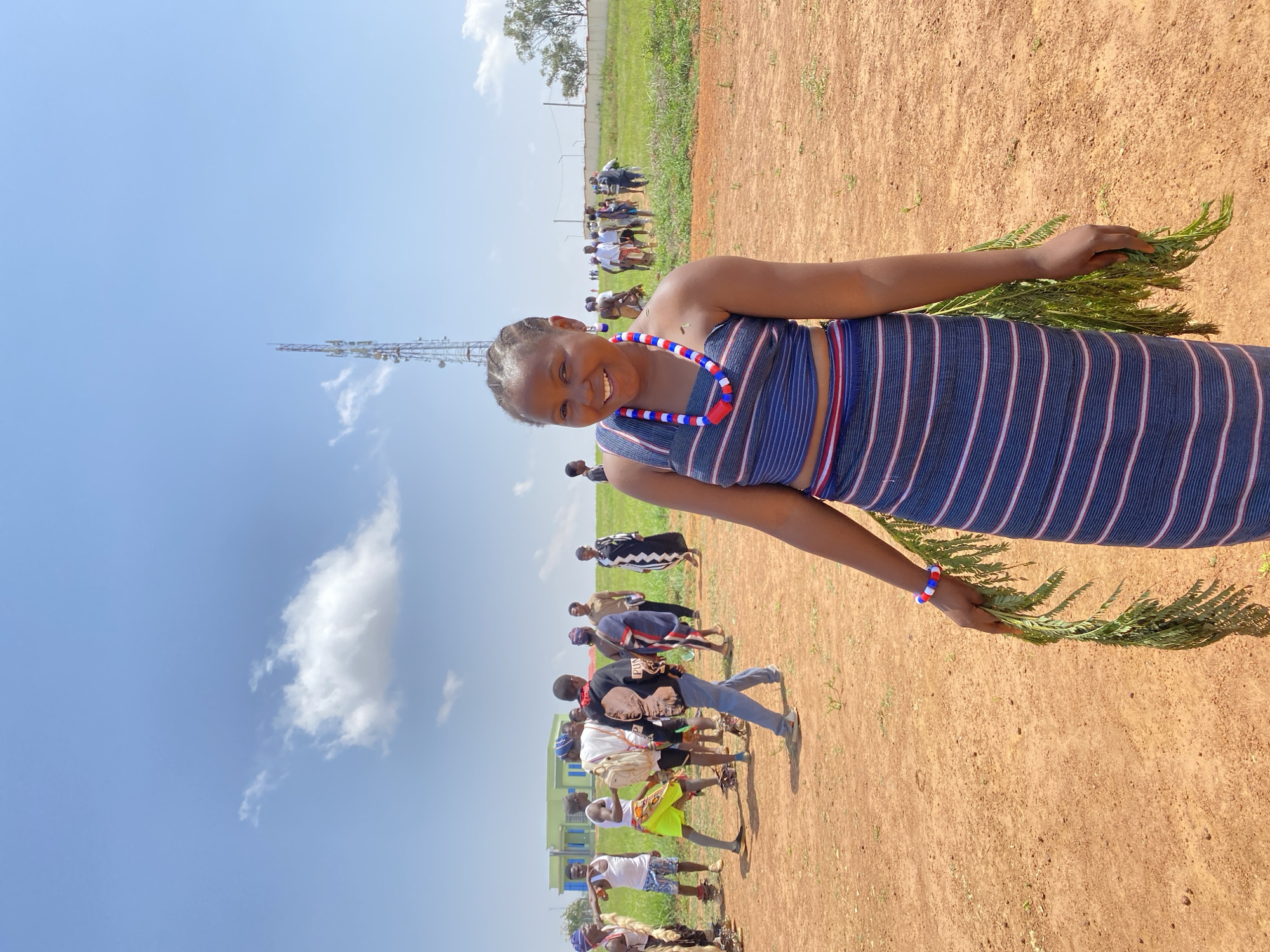 The image shows a woman in a blue and white striped traditional outfit, holding green leafy branches in her hands. She wears a red, white, and blue beaded necklace and a matching bracelet. She smiles at the camera. Behind her is a group of people, some also in traditional attire. In the background, there is a grassy field and a tall cell phone tower, under a blue sky with some clouds. The location is a field in Durumi, Nigeria.