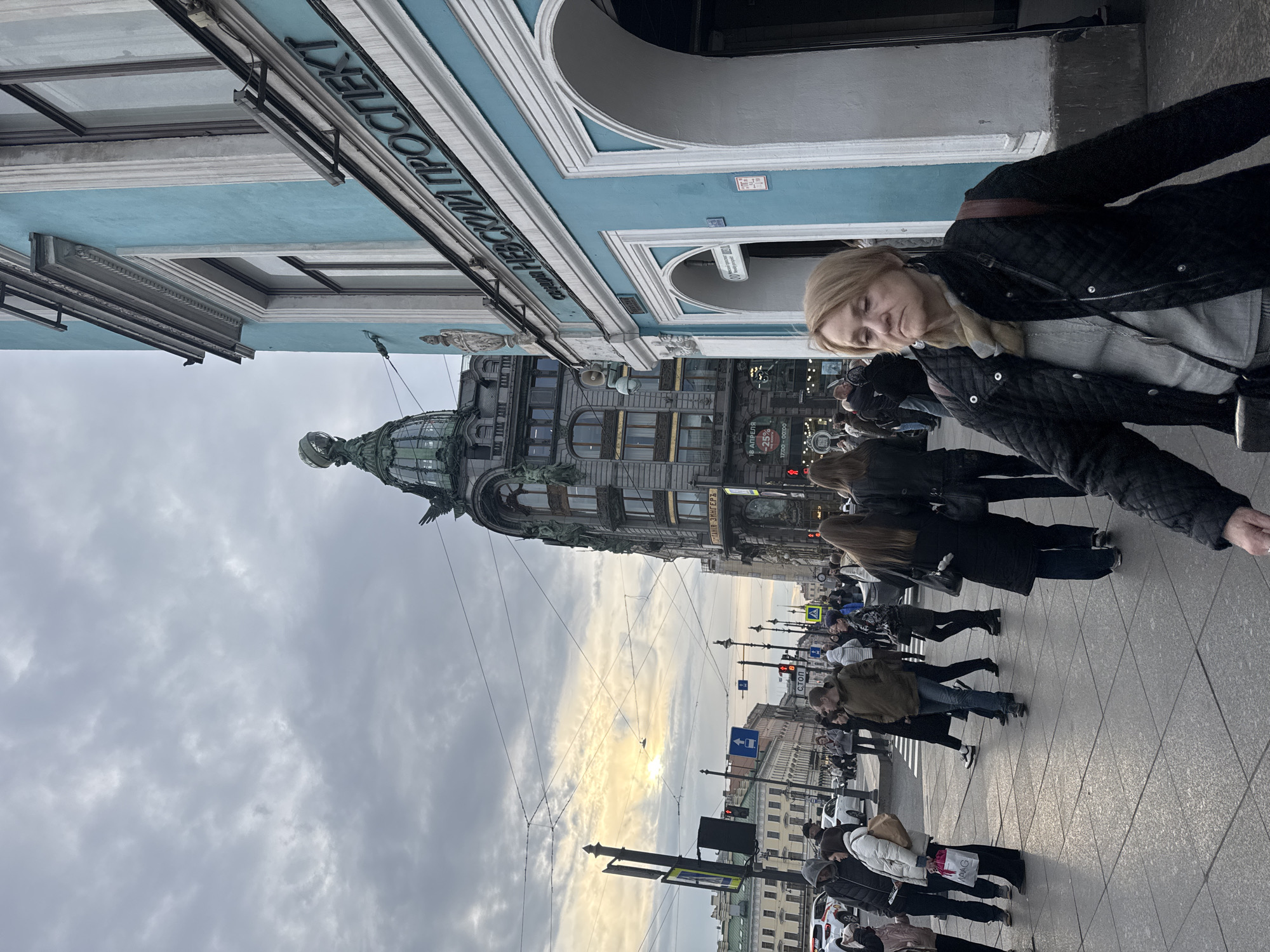 The image captures a street scene in Saint Petersburg, Russia, showcasing a bustling pedestrian environment. A woman in a black quilted jacket and a grey shirt is visible in the foreground, partially obscuring the view. The Singer House, a prominent architectural landmark with a distinctive dome, dominates the background on the left side of the frame. Several other pedestrians are visible, moving in various directions on the paved sidewalk and crosswalk. Street signs and traffic lights dot the scene, indicating an urban setting. The sky is overcast with clouds, casting a soft, diffused light over the area. The building on the upper left side has lettering in Russian that says "НЕВСКИЙ ПРОСПЕКТ," indicating Nevsky Prospect, the main street in Saint Petersburg. The signage also includes the name "ЗИНГЕРЪ" (Singer), likely referring to the Singer Company building. There are also some signs with percentages indicating a discount, and times displayed.