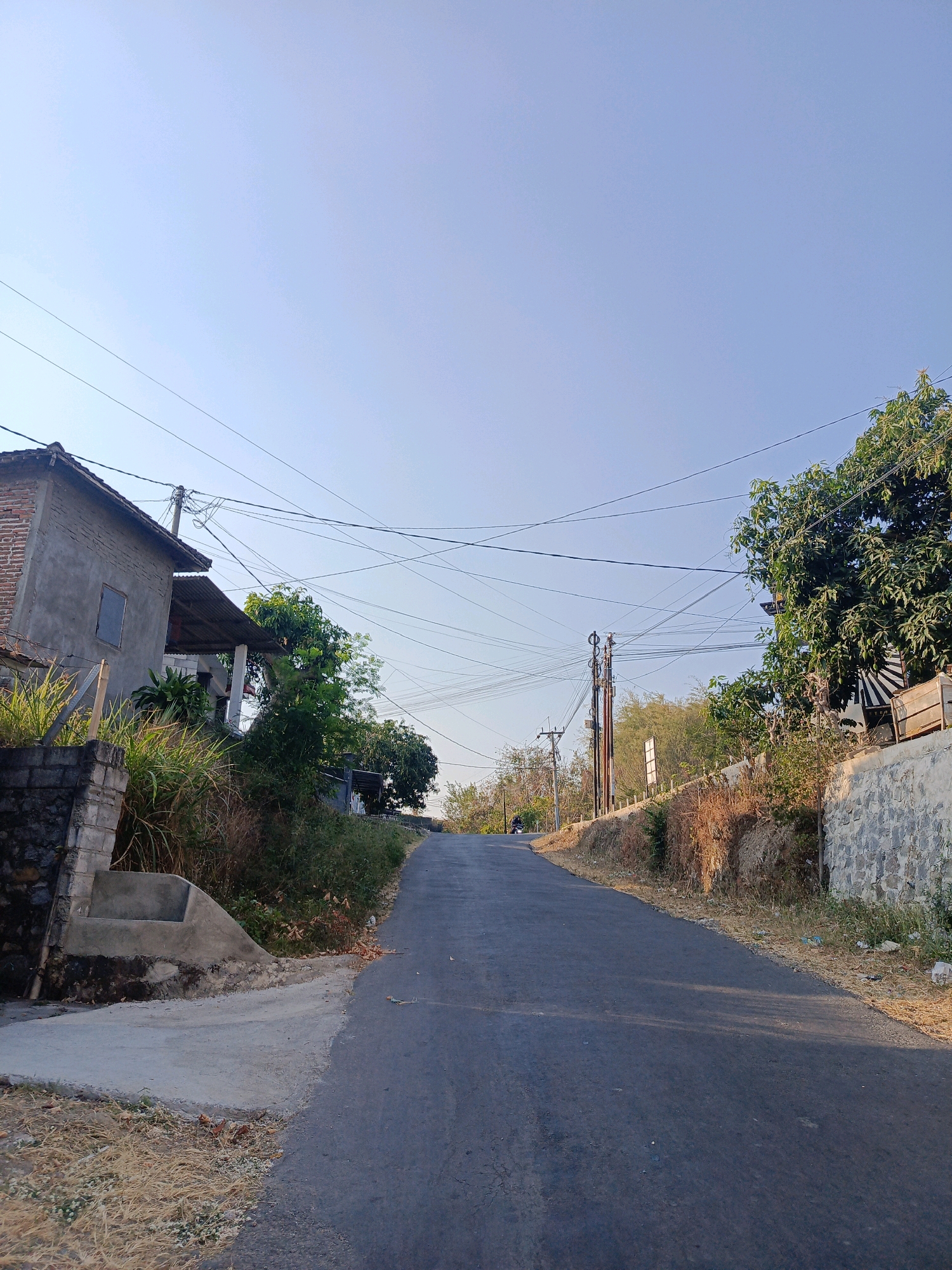 The image depicts a paved, uphill road in Ringinagung, Indonesia. On the left side of the road, a weathered, two-story house with a corrugated metal roof is visible. Adjacent to the house is a stone retaining wall and some vegetation. Overhead, a complex network of utility wires crisscrosses the sky, supported by utility poles. On the right side of the road, there is more vegetation and a stone wall. The sky is clear and blue, suggesting a daytime scene. A motorcycle can be seen in the distance on the road. The overall impression is a rural or semi-urban residential area.
