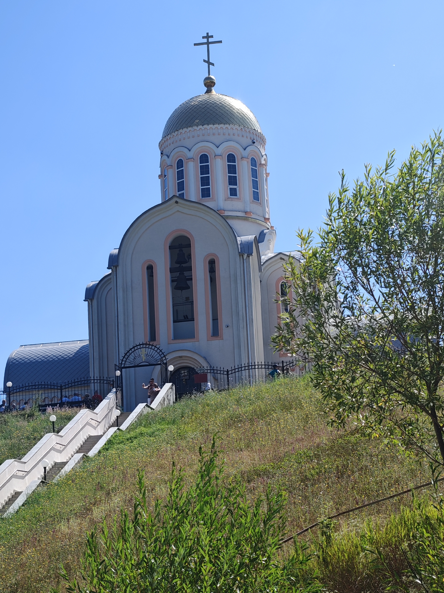 The image depicts the Cathedral of the Nativity of the Theotokos in Varvarovka, Russia, on a clear, sunny day. The architectural style is predominantly Orthodox Christian, with a prominent golden dome topped by a cross. The main structure is light-colored with arched windows and decorative pink trim. A grand staircase, lined with lampposts, ascends a grassy hill towards the cathedral's entrance, which is partially obscured by a black metal fence. Several people are visible on the stairs and near the entrance. Lush green foliage, including trees and overgrown grass, surrounds the scene, suggesting a natural, possibly semi-wild environment. The sky is a vibrant, cloudless blue.