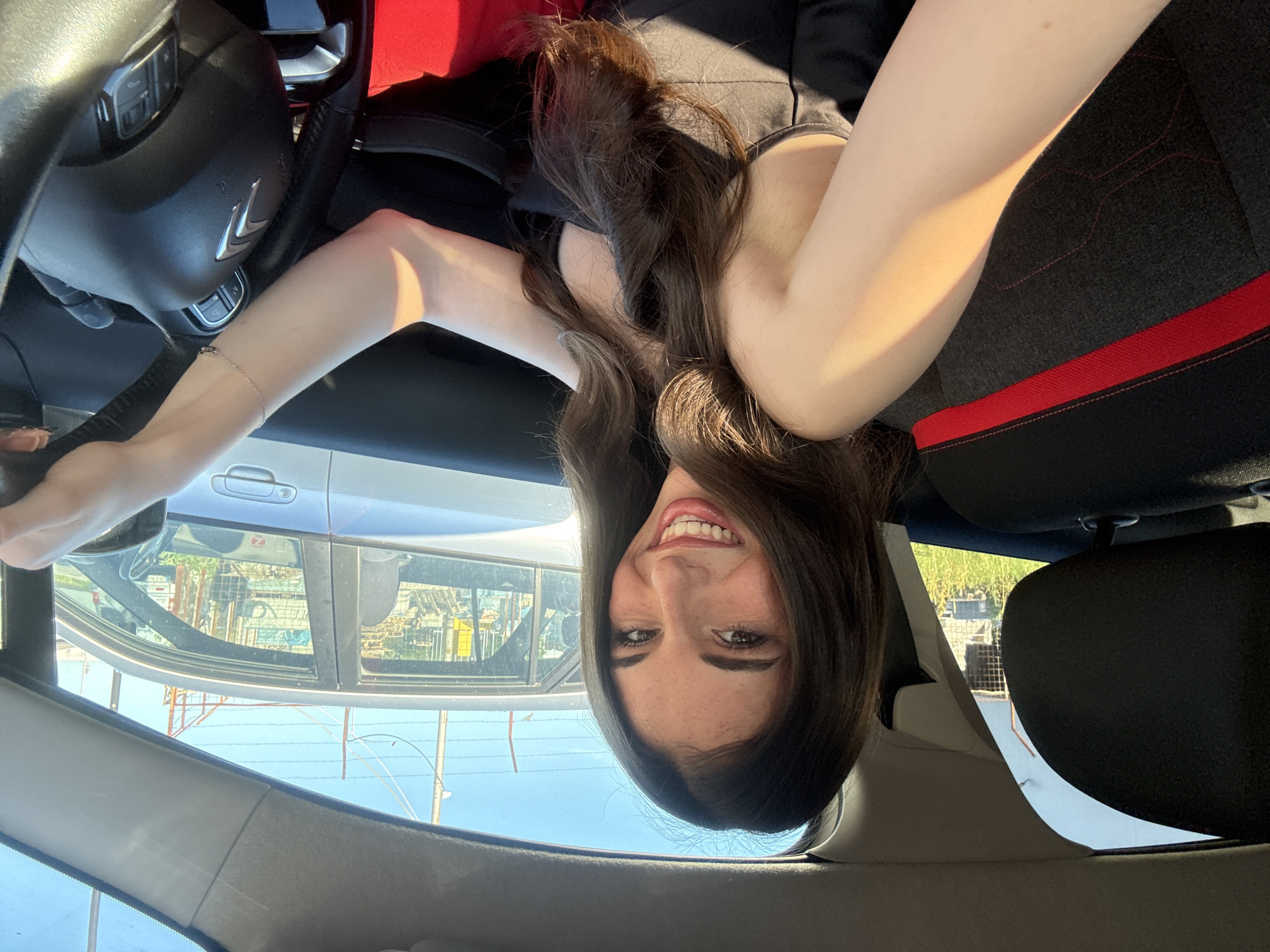 A woman is inside a car. The woman has brown hair and is smiling. She is wearing a black top. The car has a black steering wheel with a silver logo. The seats are black with red trim. Outside the car, there are buildings and power lines. The photo appears to be taken upside down. Based on the style of buildings, the environment visible through the car window, and the request, the general location may be Plovdiv, Bulgaria.