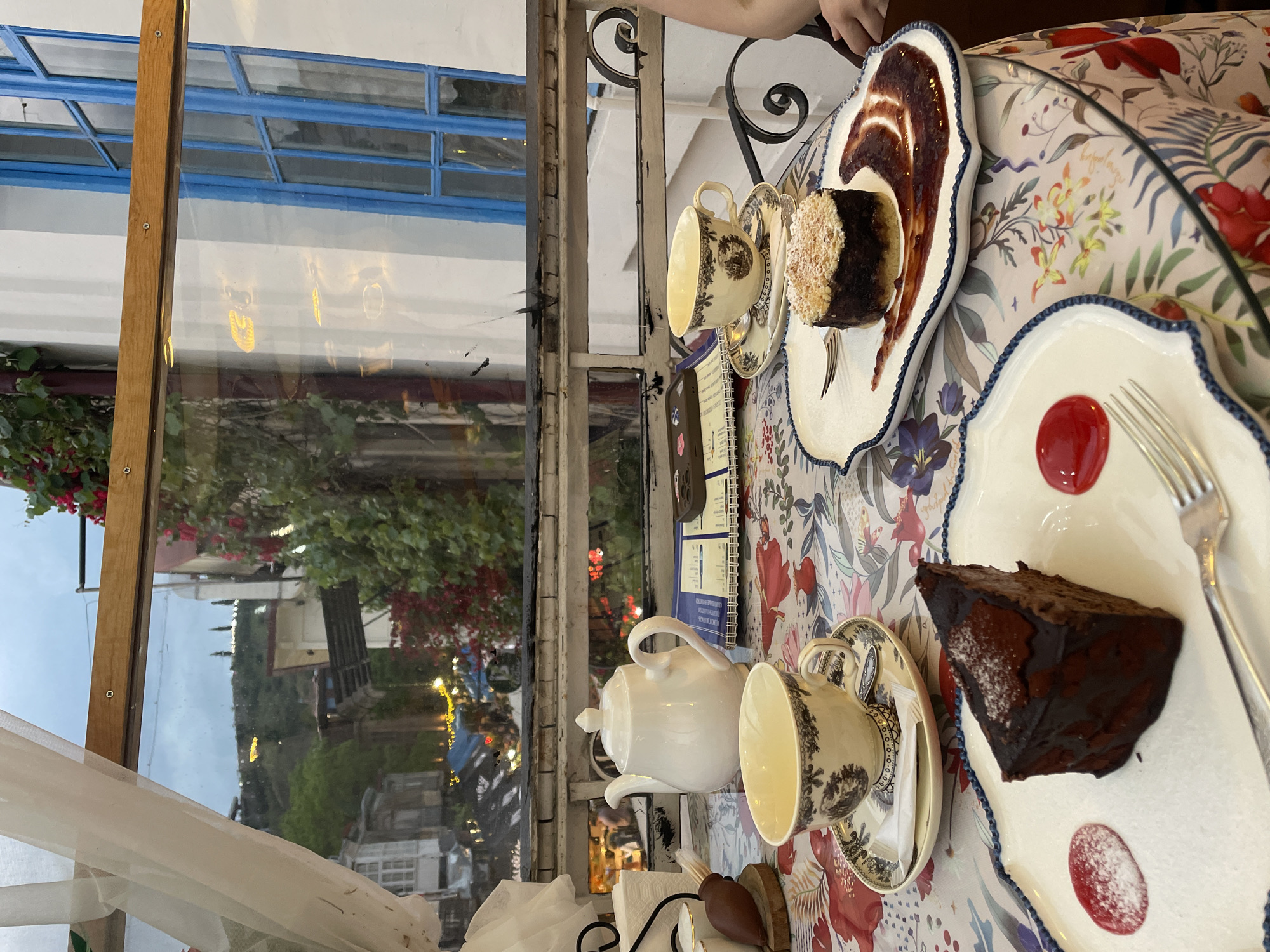 The image shows a table setting at a cafe, likely in Tbilisi, Georgia, given the reflection in the window of the city's characteristic architecture and lighting. The table is covered with a floral-patterned tablecloth and set with two ornate plates, each holding a slice of cake and adorned with a dollop of red sauce. A fork rests on one plate. Two matching teacups sit on saucers, one near a teapot. The reflection in the window reveals a street scene with trees, buildings, and streetlights, suggesting an outdoor cafe setting or a view from an interior location. The scene is captured from a high-angle perspective, offering a view of the table setting and the surrounding environment.