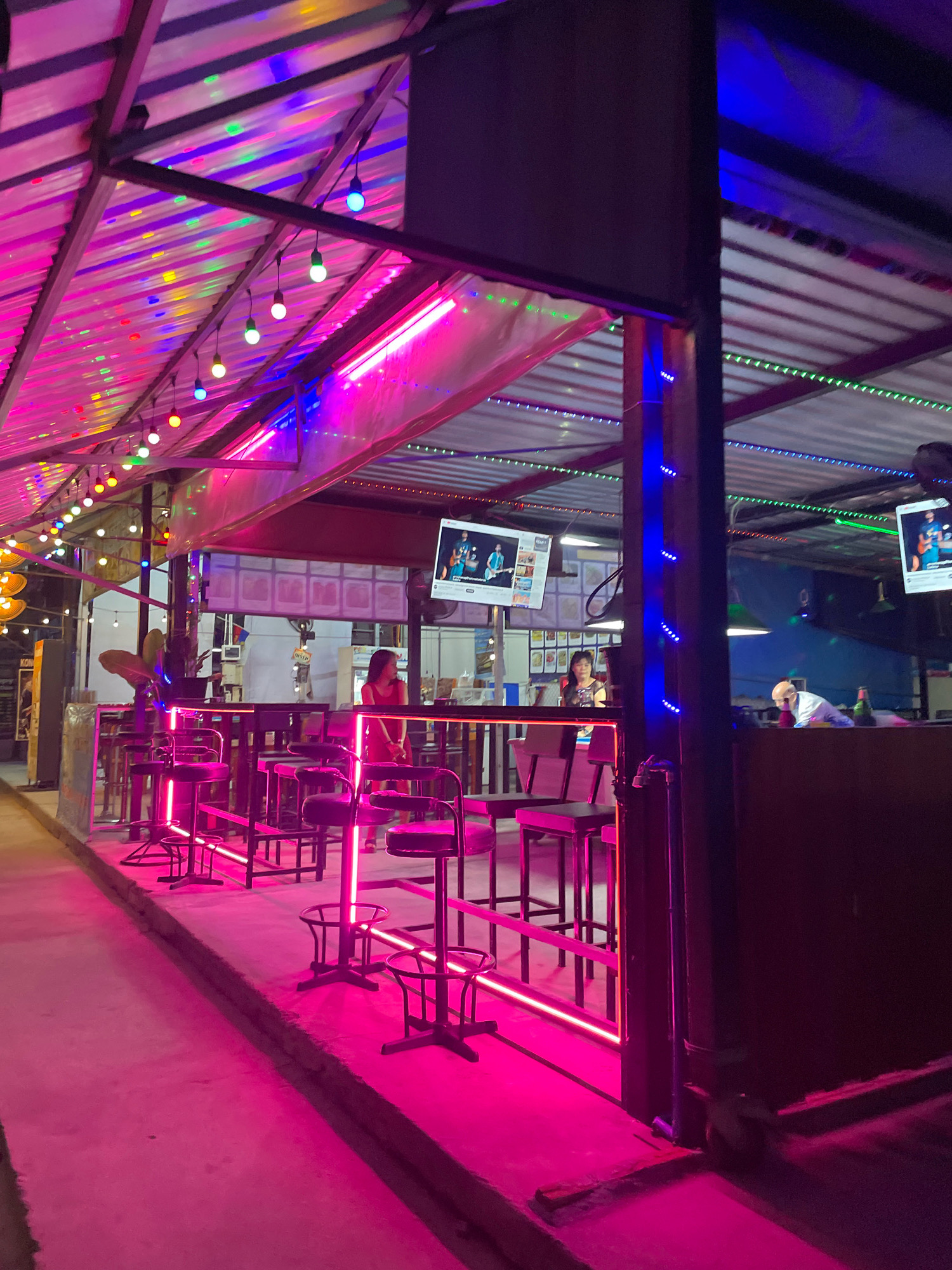 The image depicts an outdoor seating area of a bar or restaurant in Pattaya City, Thailand, at night. The area is illuminated with vibrant pink and blue neon lights, and strings of colorful bulbs hang from the corrugated metal roof. Several high-top tables with stools are set up, with some of the stools and table bases featuring pink neon lighting. In the background, two women are visible. One woman in a red dress stands near a table, while another woman with dark hair is seated at a bar. A television screen displaying an image of people is mounted on the wall. The ground is paved and reflects the pink lighting. The overall atmosphere suggests a lively nightlife scene.