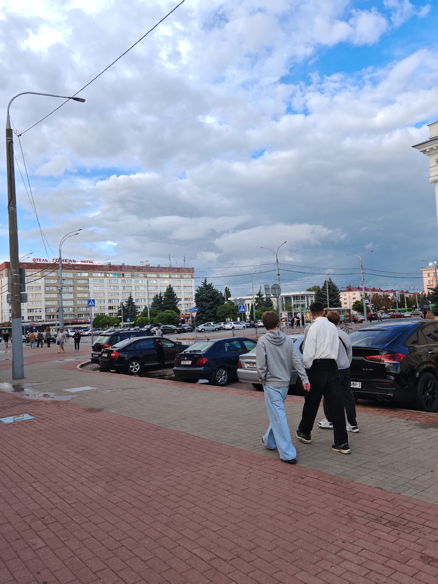 The image depicts a street scene in Homyel, Belarus, with a large hotel building in the background. The hotel, identified by the sign "ОТЕЛЬ ГОМЕЛЬ HOTEL," is multi-story with numerous windows. In the foreground, a paved walkway made of red and gray bricks is visible, with some puddles on the ground, suggesting recent rain. Several cars are parked along the street, including a black Mercedes-Benz SUV with "GLE 53" visible on the back.
Three young men are walking away from the camera on the brick walkway. The person in the middle wears a white shirt and black pants, while the person on the left wears a gray hoodie and light blue jeans. The person on the right also wears a gray hoodie and dark pants. In the midground, other pedestrians are walking along the sidewalk in front of the hotel. The sky is partly cloudy with a mix of blue sky and large, gray clouds. Trees and streetlights are also present in the scene. The overall impression is an urban environment on a cloudy day.