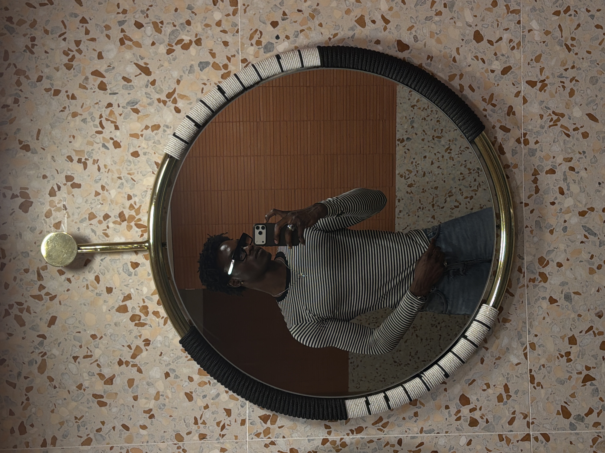The image shows a selfie taken in a bathroom with speckled tan and grey stone tile walls, and a reflection of brown, stacked tile. The subject, a person with dark hair and skin wearing dark glasses and a black-and-white striped long-sleeved shirt and jeans, is reflected in a round mirror with a brass frame. The mirror's frame is wrapped with sections of woven white and black material. A brass cylindrical fixture extends horizontally from the left side of the mirror. The subject is holding a smartphone to take the selfie. The specific location in London, United Kingdom, cannot be determined based on the image alone.