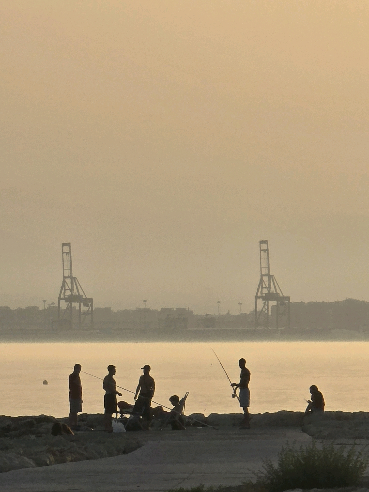 In a coastal area of Málaga, Spain, several individuals are silhouetted against the hazy, golden light of a sunrise or sunset. The scene depicts a group of people fishing from a rocky shoreline, with a body of calm water in the foreground. In the background, a distant cityscape is visible, characterized by industrial cranes and buildings, suggesting a port or industrial zone. The atmosphere is serene, with the soft light casting long shadows and creating a muted color palette dominated by warm tones. The people are engaged in various activities, including holding fishing rods, sitting, and conversing, while the environmental elements contribute to a sense of tranquility and vastness.