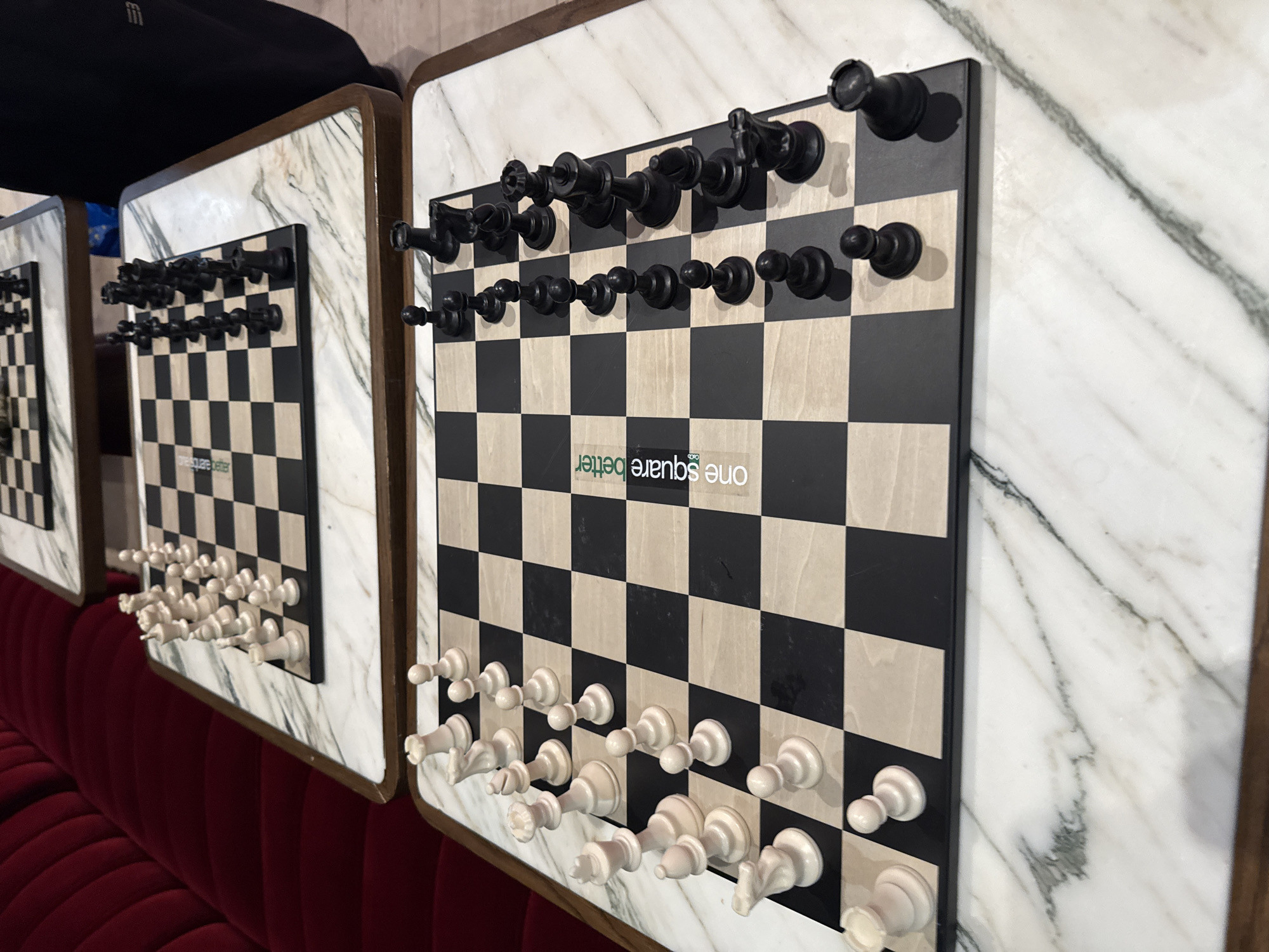 The image shows three vertical chessboards hanging on a wall. The chessboards are made of black and white squares and have the pieces attached to them. The chessboards are framed with a marble-like material. Below the chessboards is a red velvet couch. The chessboards have the words "one square better" on them. The location is unknown. The pieces are arranged in the traditional starting positions for a game of chess.