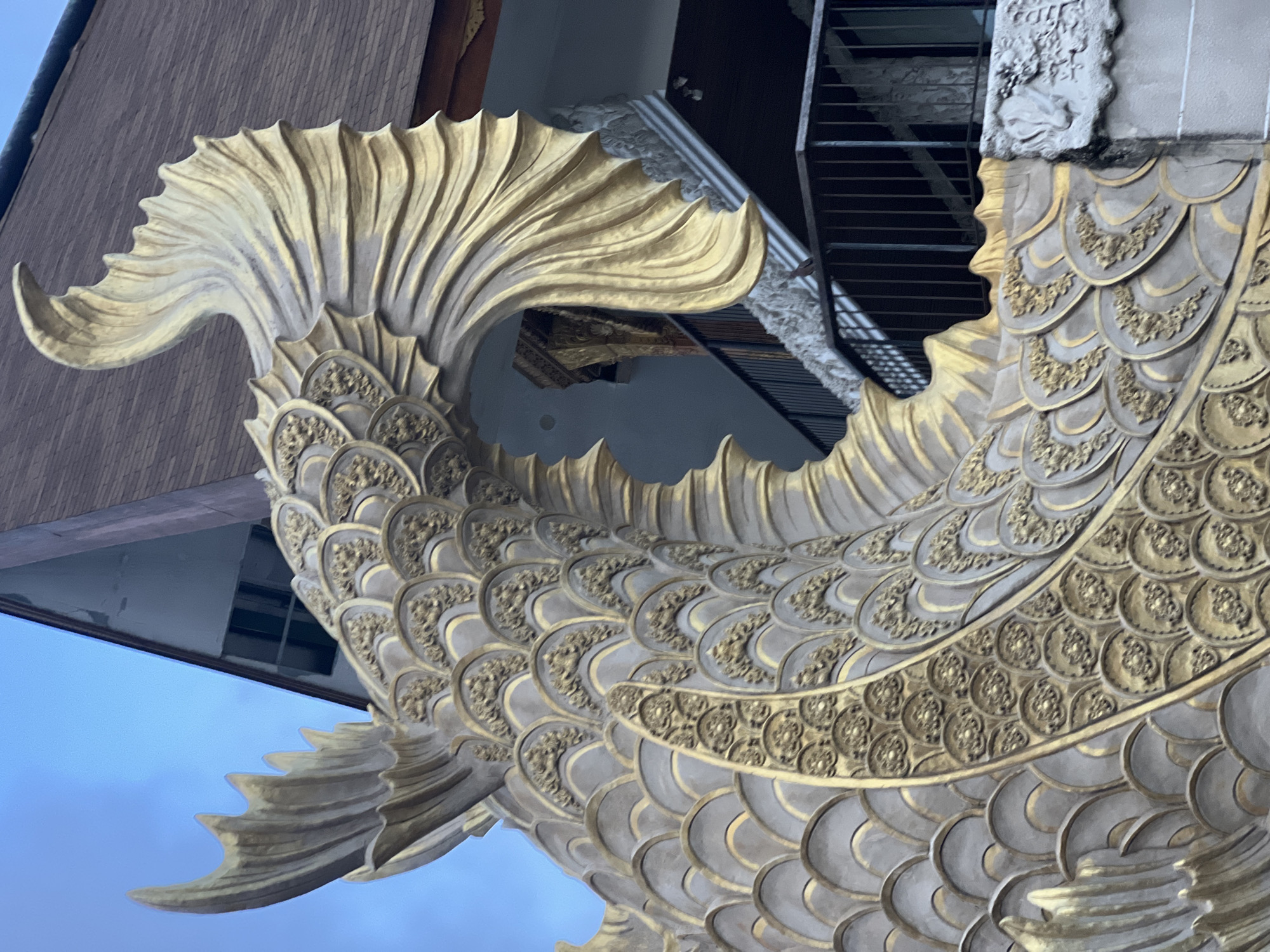 The image displays a close-up of a large, ornate sculpture of a mythical fish or serpent-like creature, possibly a Nāga, integrated into a building structure. The sculpture is rendered in shades of gold and light gray-gold, with intricate detailing. Its body is covered in layered scales, where the outer edges are a lighter tone, and the inner, raised patterns within each scale are a brighter, more lustrous gold. A prominent decorative band with circular golden motifs runs horizontally across the creature's mid-section. The large, flowing tail and several fins are also highly stylized, featuring wavy, pleated textures and golden hues, suggesting motion.
The sculpture appears to be part of an architectural facade, with elements of the building visible in the background. To the upper left, there's a dark, wooden-paneled surface, possibly siding. To the upper right, concrete structures with some carved details and dark metal stairs or railings are visible. The sky, seen in gaps, is a clear, light blue, indicating a daytime setting. No people or specific activities are present. The elaborate craftsmanship and mythical subject matter are consistent with traditional Balinese artistry, fitting its location in Denpasar, Indonesia. No discernible text is visible anywhere in the image.