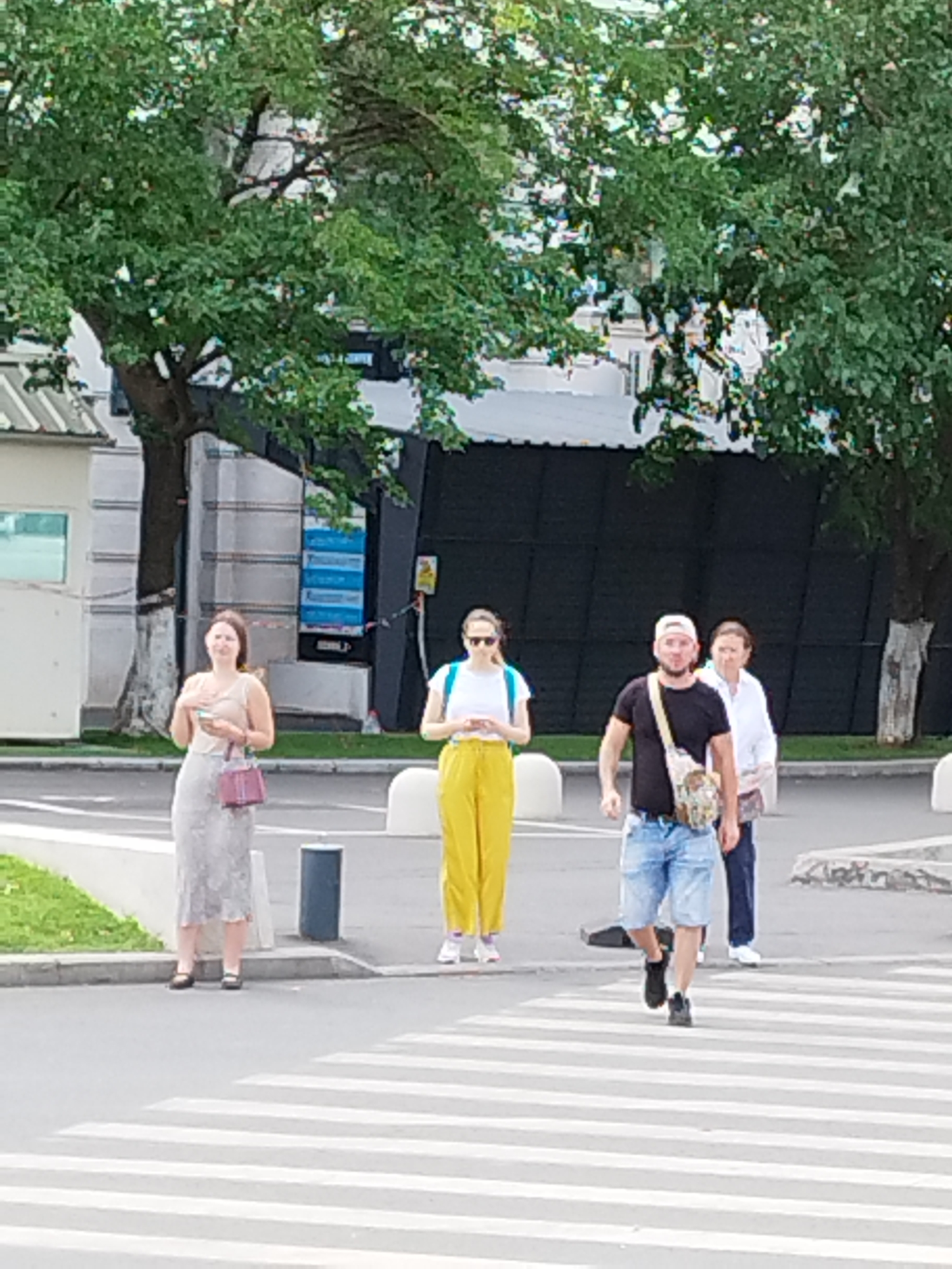 The image captures a street scene in Bucharest, Romania, featuring four individuals near a crosswalk. In the foreground, a crosswalk with white stripes is visible on asphalt. To the left, a woman stands wearing a light-colored dress and holding a maroon handbag. Next to her, a woman in yellow pants and a white shirt with a blue backpack is also standing. Further to the right, a man in a black t-shirt, blue shorts, and a white baseball cap, with a patterned bag slung across his body, walks across the crosswalk. Behind him, a woman in a white shirt and dark pants walks. The background consists of trees, buildings, and a dark fence. The lighting suggests it is daytime.