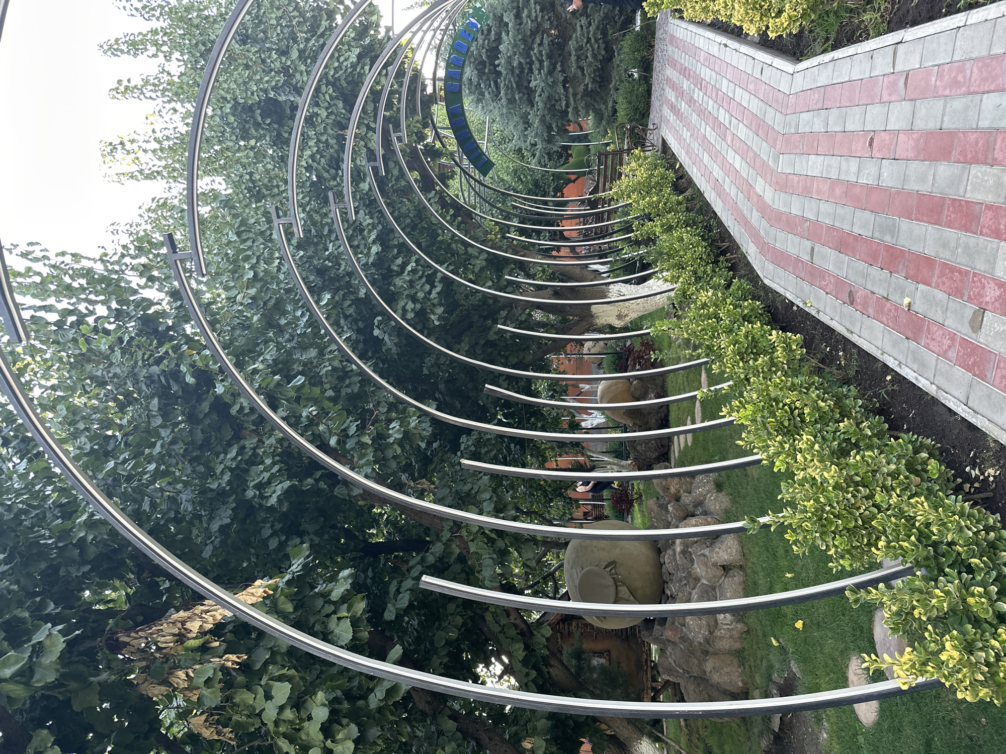This is a photograph of an arched walkway in Parakar, Armenia, likely within a garden or park. The walkway is paved with red and gray rectangular stones arranged in a diagonal pattern. Overhanging the walkway are metal arches with white accents. Lush green bushes line the walkway on the left side, and a line of similar bushes is also visible on the right. In the background, mature trees with dense green foliage create a canopy. To the left, there's a large, spherical stone sculpture with a brown covering. Further back, a small rustic wooden structure is visible. To the right, a conifer tree stands tall, and in the distance, a green banner with text, possibly "MIRAMANA GARDEN", is partially visible. A park bench is situated along the walkway. The sky appears overcast.