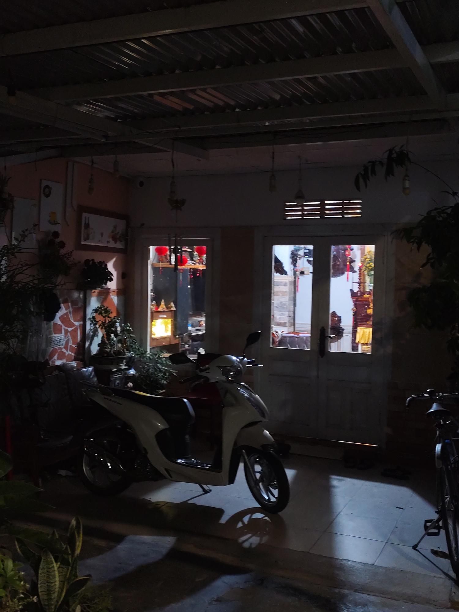 The image depicts an outdoor covered area, possibly a porch or carport, at night. A white and red scooter is parked in the foreground, casting a shadow on the tiled floor. To the right, the front of a bicycle is visible. The background shows a doorway with glass panels, revealing an interior space with decorative elements and what appears to be an altar. The ceiling is corrugated metal supported by wooden beams, and several bare light bulbs hang from wires. The overall atmosphere is dimly lit and somewhat cluttered, with plants visible in the foreground and to the sides. The scene does not provide explicit visual cues to pinpoint a specific location in Da Lat, Vietnam, but it suggests a residential or small business setting.