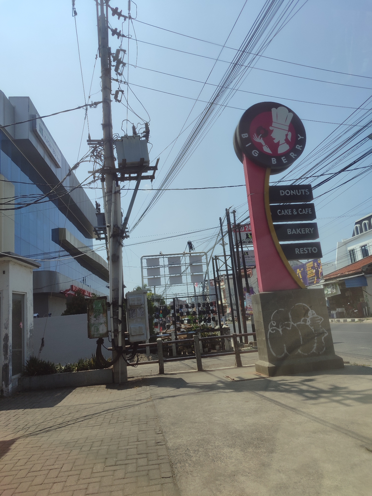 This is a photograph of a street scene in Tegal, Indonesia. The image features a large, modern building with a glass facade on the left, partially obscured by a utility pole with a transformer and numerous electrical wires. To the right of the pole is a tall, curved sign for "BIG BERRY," a business that offers donuts, cake & cafe, bakery, and resto services. The sign is predominantly pink and yellow and has a circular logo at the top depicting a chef's hat and profile. Below the logo, the sign lists its services. A sidewalk paved with interlocking bricks is in the foreground, leading to a concrete street. The sky is clear and blue, indicating a sunny day. The background shows more buildings, some with arched roofs, and various street-level businesses. Graffiti is visible on the base of the Big Berry sign. The scene is typical of a commercial area in an Indonesian city.