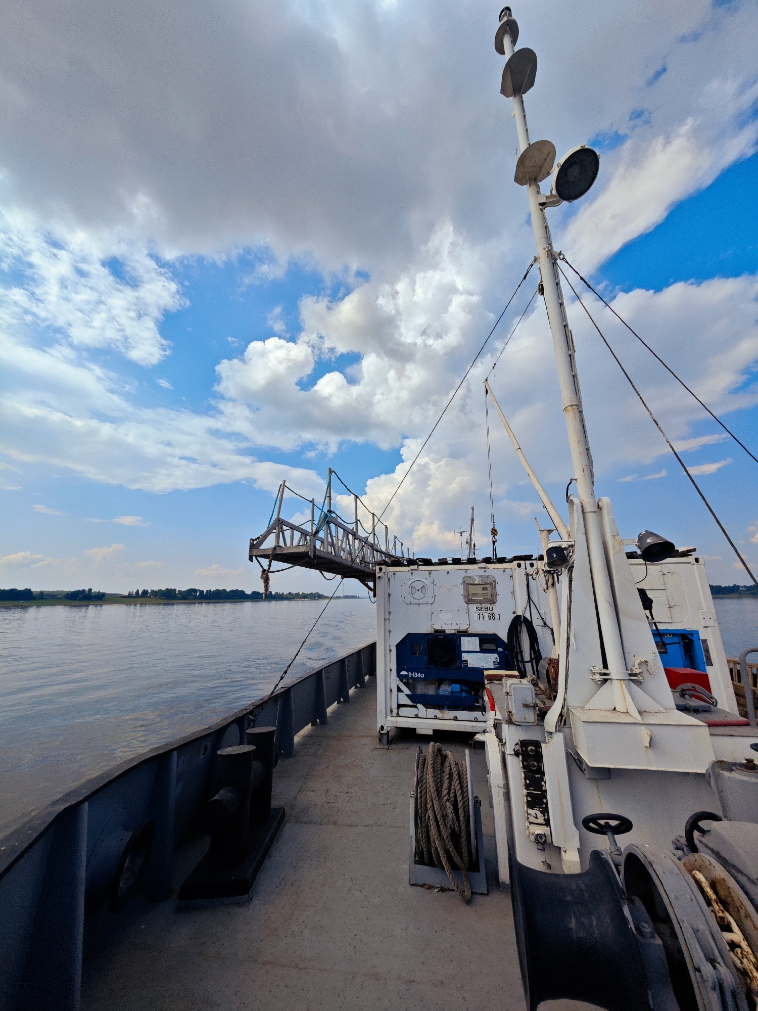 The image depicts the deck of a vessel navigating on the river in Erkalovo, Russia. The perspective is from the stern looking forward and to the port side. A metal railing runs along the left edge of the deck, with a large black bollard mounted on it. The water of the river is calm and extends to the horizon, where a treeline is visible. Above, a dramatic sky filled with white and gray clouds is present.
Dominating the right side of the frame is the vessel's superstructure, painted white. A tall mast with multiple lights and sensors extends upwards. Various equipment is visible, including a white container with markings such as "SEBU 11 68 1" and what appears to be a blue refrigeration unit labeled "R-134a". A coiled rope rests on a stand to the left of the container. Towards the foreground, a winch mechanism is partially visible. A gangway structure, with ropes and a railing, extends from the vessel towards the viewer's left, suggesting it might be deployed or ready for use. No people are visible in the image.