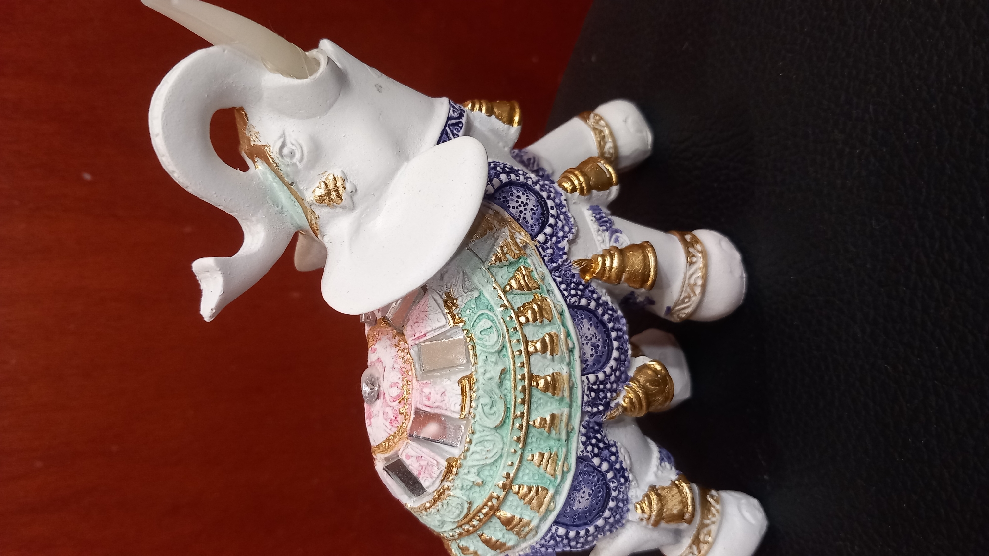 A white, decorative ceramic elephant figurine with an elaborate saddle is positioned against a plain, dark red background. The elephant is facing to the right, with its trunk curled upwards. The saddle is adorned with colorful patterns, gold accents, small mirrors, and numerous gold bells hanging from its edges. The elephant's legs are also decorated with gold bands. The figurine is placed on a dark, textured surface, possibly a table or chair. The scene is well-lit, highlighting the details of the figurine. There is no information about the location in the city of Kos, Greece.