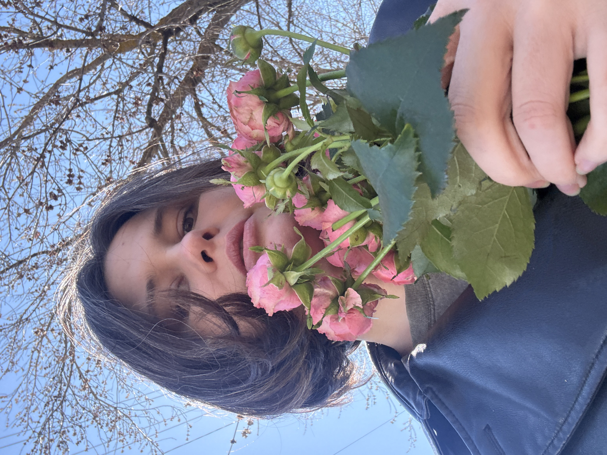A low-angle, rotated close-up captures a person holding a bunch of small pink roses with green leaves and buds, partially obscuring their face. The person, with dark brown hair, looks directly towards the camera with a neutral expression. Only one eye, the nose, and the lips are clearly visible. They are wearing a dark, possibly leather, jacket over a grey garment.
The background features a clear blue sky with numerous bare tree branches silhouetted against it, indicating a setting in late winter or early spring. The lighting is bright and natural, suggesting it is daytime. The overall scene conveys a casual outdoor portrait in Фоки, Russia. No text is visible in the image.