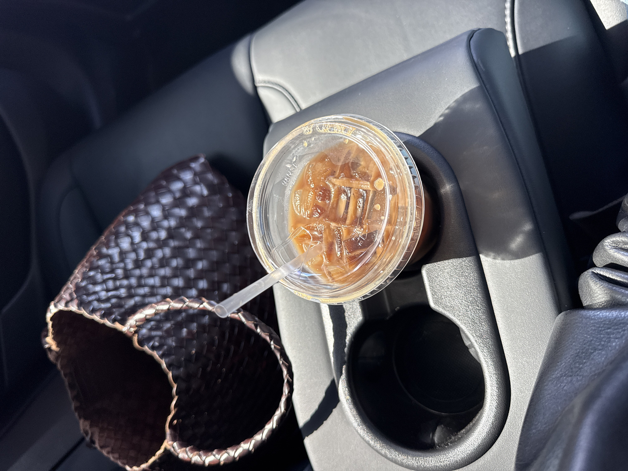 The image shows a car interior. There is a cup of iced coffee with a straw in the cupholder. Beside the cupholder is a brown, woven bag. The car seats are black leather. This is likely in Los Angeles, United States of America.