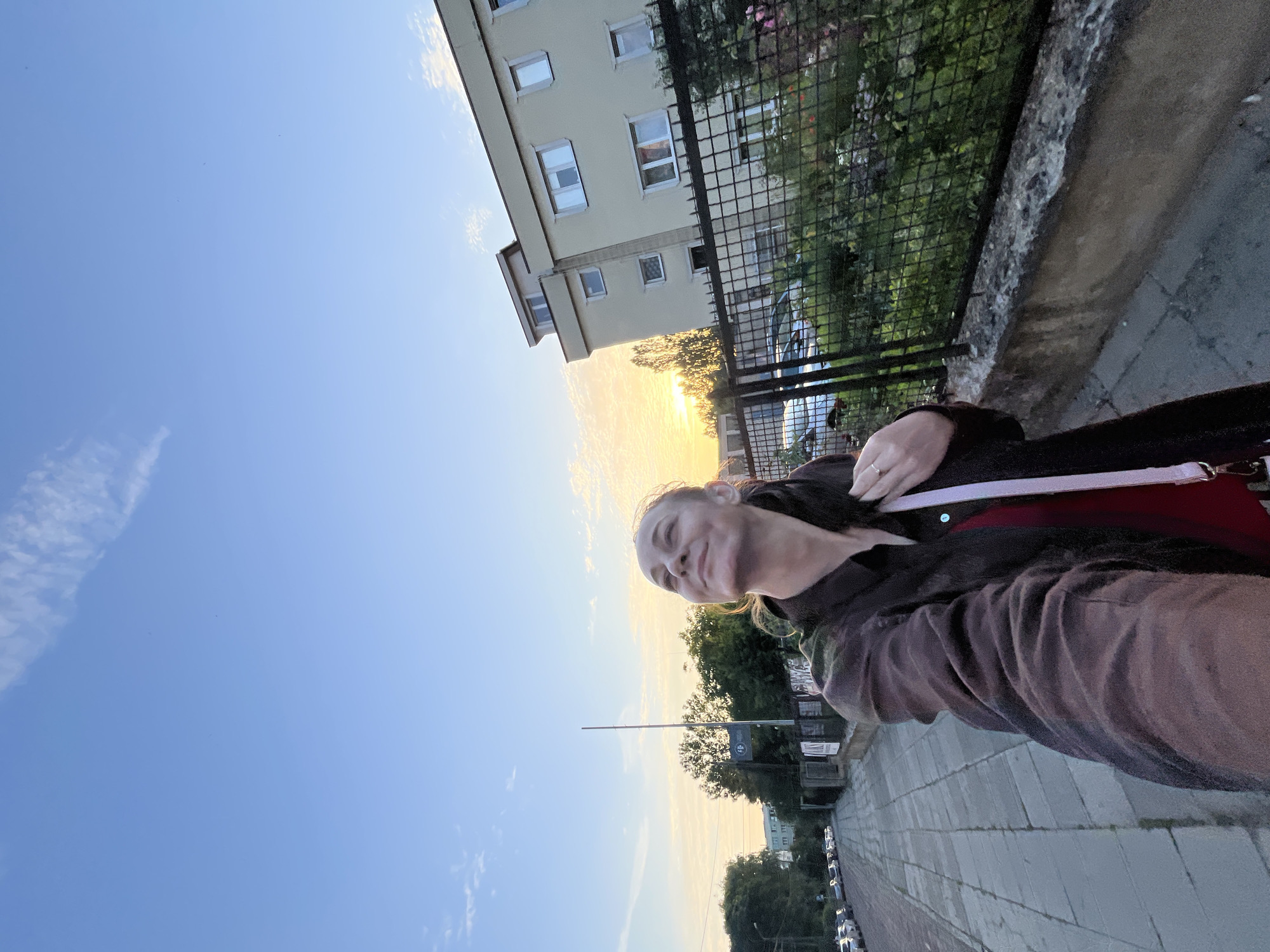 The image shows a woman taking a selfie outdoors. The woman is smiling and looking to her left. She is wearing a dark brown jacket over a red top. A ring is visible on her left hand. The setting appears to be a sidewalk next to a building with a black fence in front of it. Trees and cars are visible behind the fence. The sky is blue with some scattered clouds, and the sun is setting, casting a warm glow on the scene. The overall mood is relaxed and pleasant.