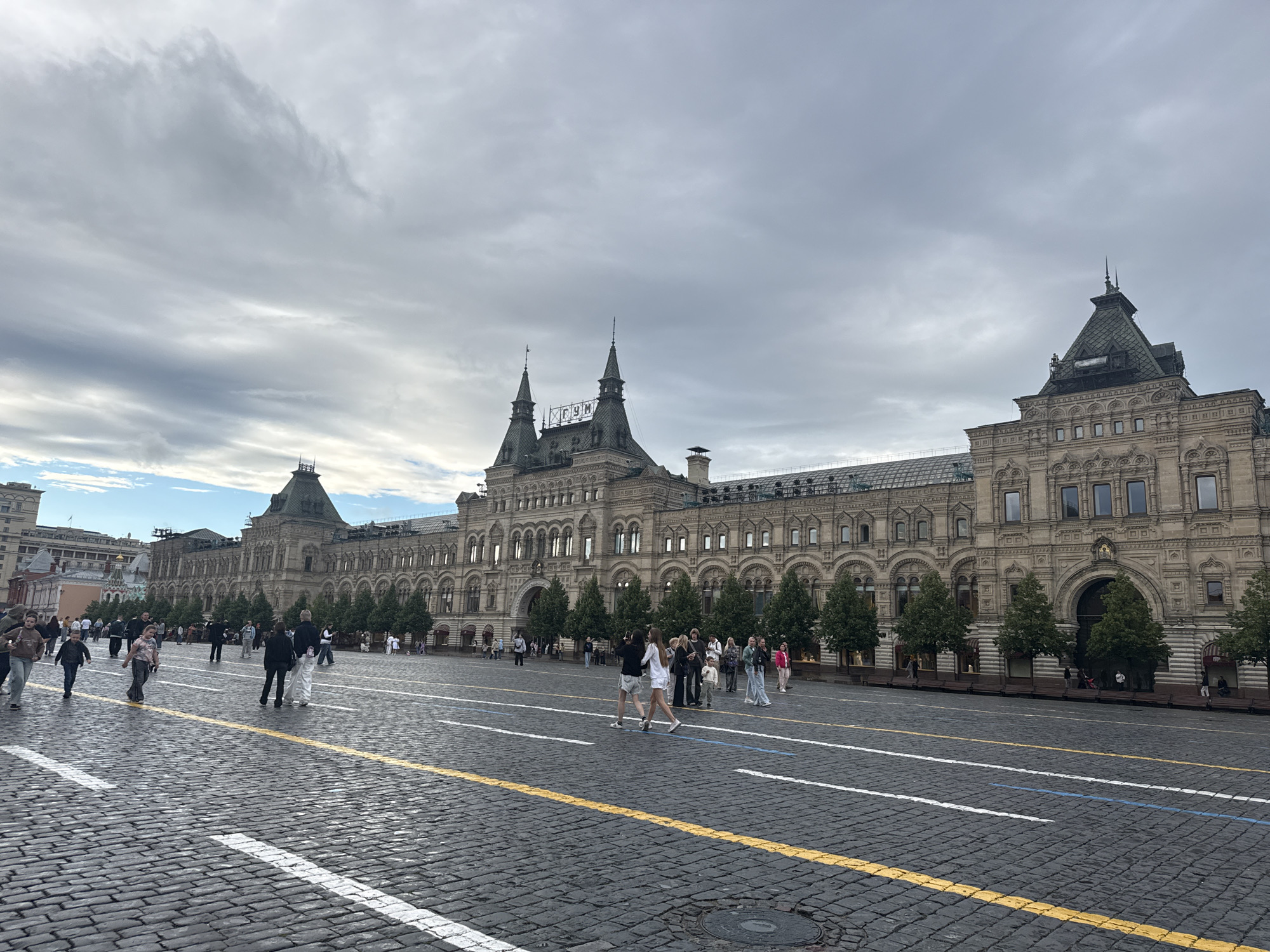 The image depicts a wide, cobblestone plaza in front of a large, ornate building with a distinctive roofline and multiple spires. Several people are scattered across the plaza, some walking, others standing in small groups. The building appears to be a historic architectural landmark, possibly a department store or a government building, with many windows and decorative elements. Trees line the front of the building, and there are benches in front of them. The sky is overcast with clouds, suggesting a cloudy day. The lighting is diffused, and there are no harsh shadows, indicating it is likely daytime, though not necessarily bright sunlight. The overall atmosphere seems calm and public, with individuals going about their day in a tourist or public space. The cobblestone paving and the grand architecture are key visual elements. There are white and yellow lines painted on the cobblestones, indicating a designated path or area. The building has a sign with Cyrillic letters visible at the top of one of its central spires, reading "ГУМ".